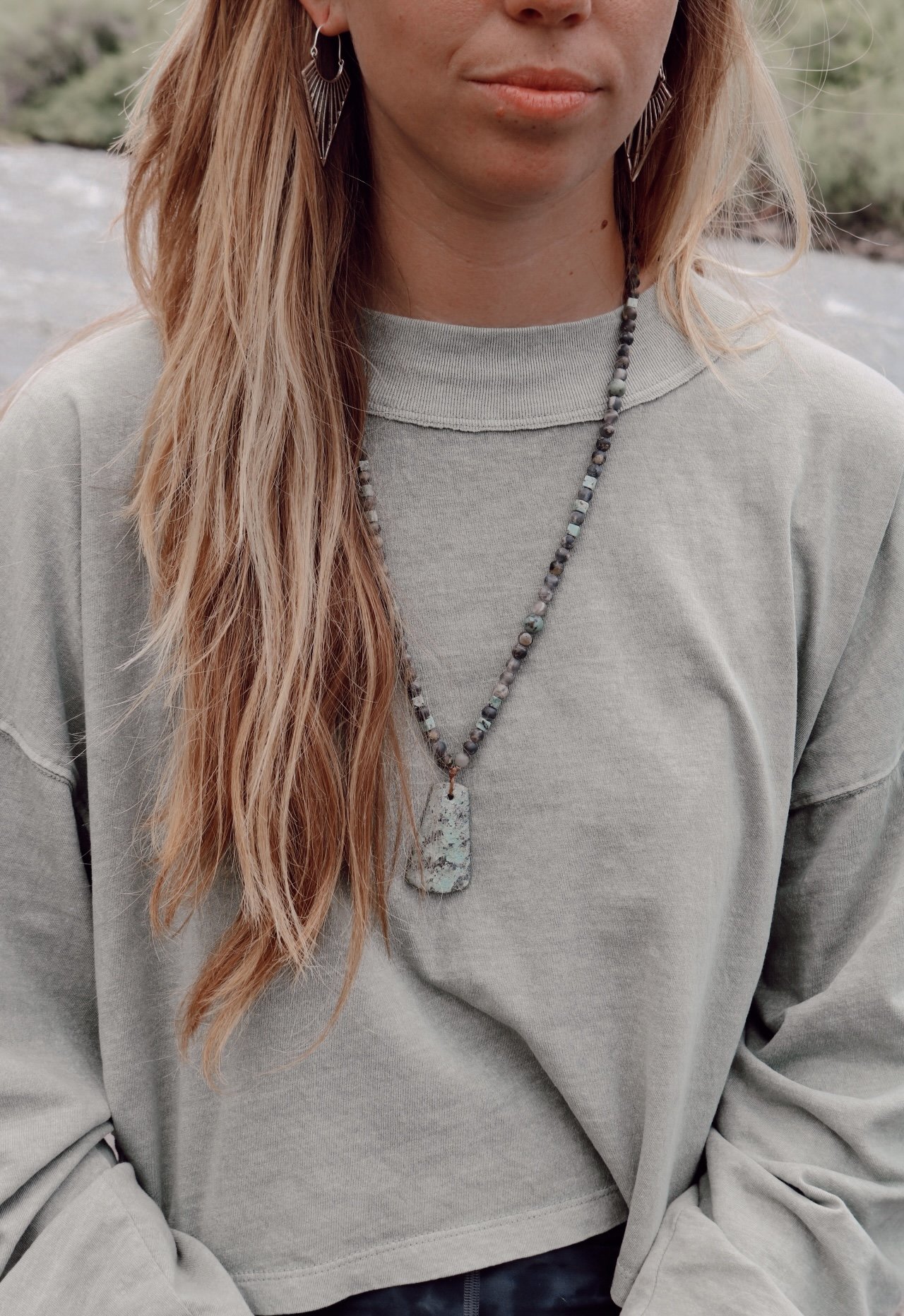 A woman with long, wavy, reddish-blonde hair, wearing earrings and a beaded necklace with a large, rectangular stone pendant, dressed in a gray sweatshirt, standing outdoors near a river or stream.