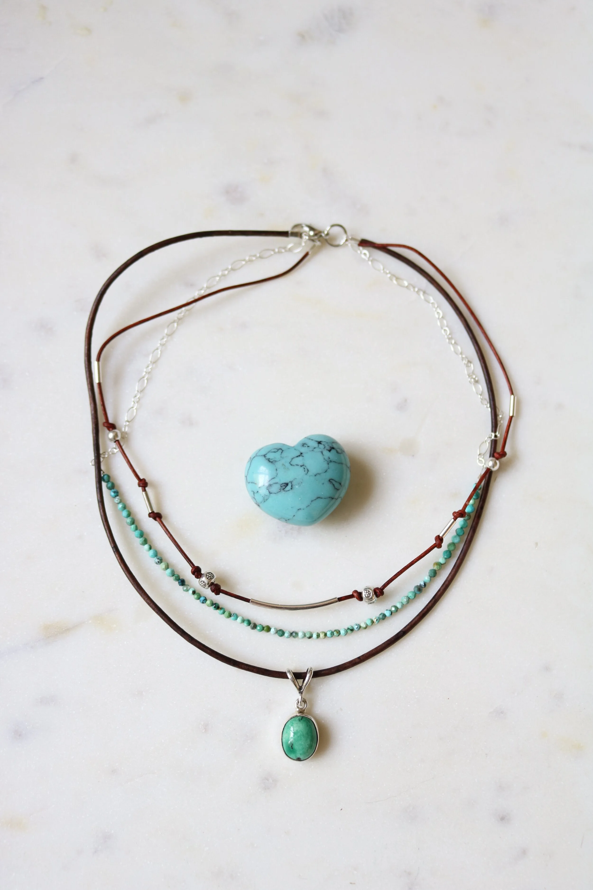 Three layered necklaces with beads and pendants, a turquoise heart-shaped stone in the center, on a white marble surface.
