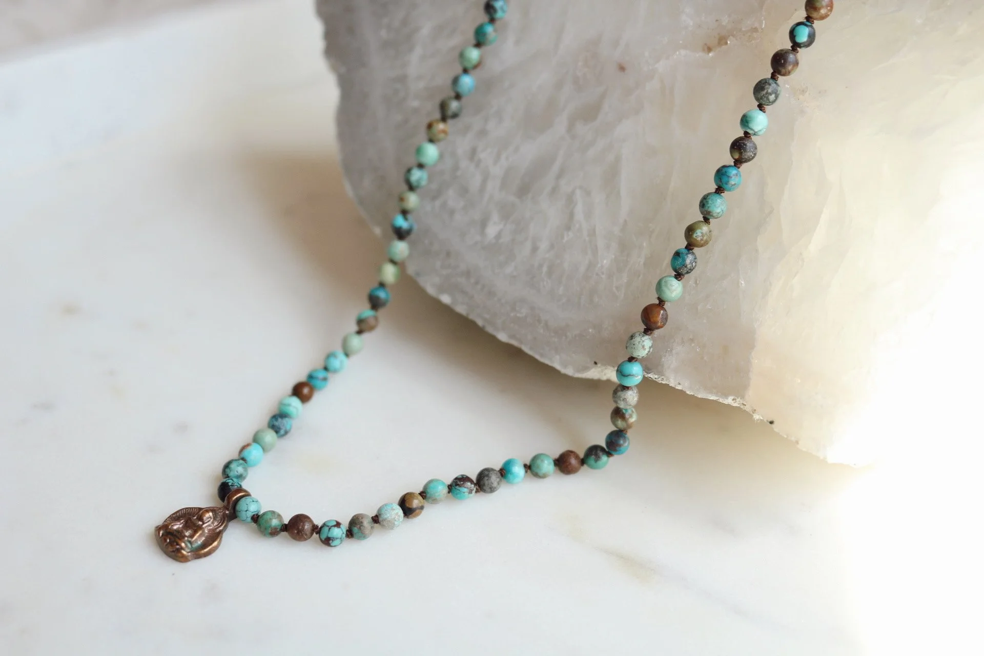 A beaded necklace with a bronze pendant resting on a white surface next to a large piece of translucent mineral or crystal.
