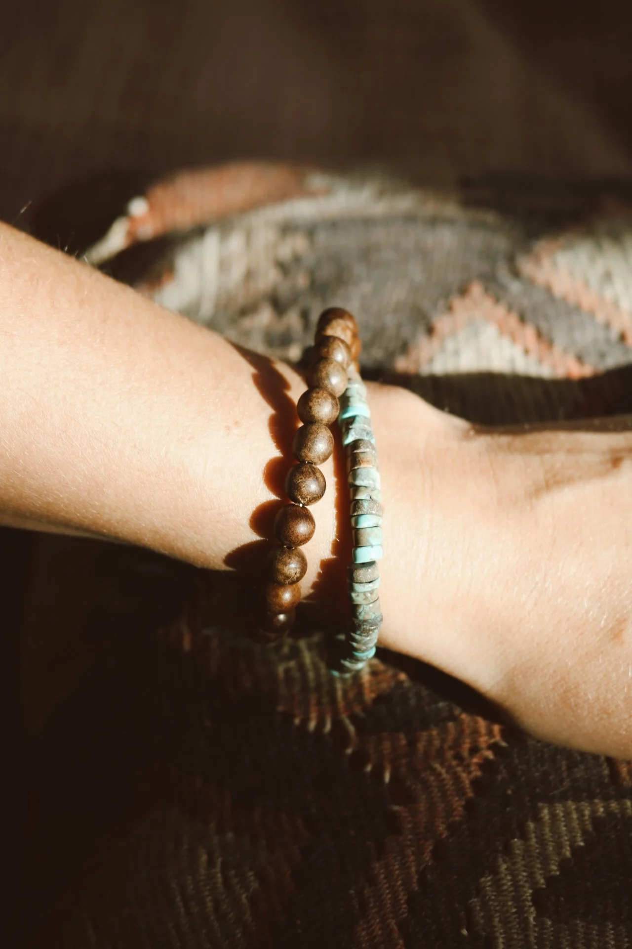 Two beaded bracelets, one with brown round beads and the other with flat turquoise beads, worn on a person's wrist. The person is wearing patterned clothing and the photo is taken with sunlight.