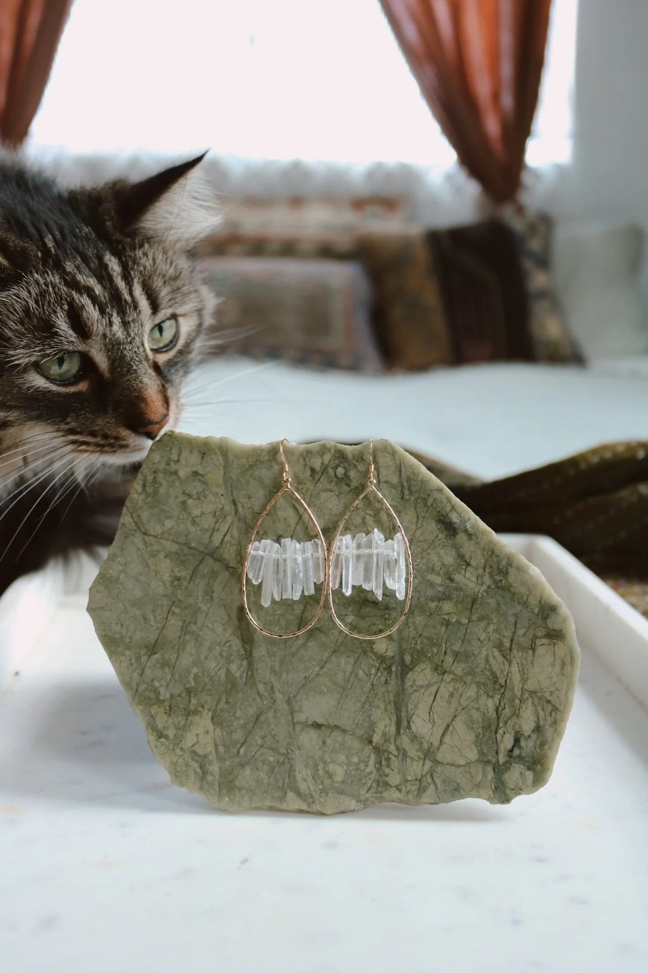 Close-up of a tabby cat sniffing a pair of gold teardrop-shaped earrings with white crystal accents, resting on a large, rough green stone on a white surface, with a cozy, blurred background.