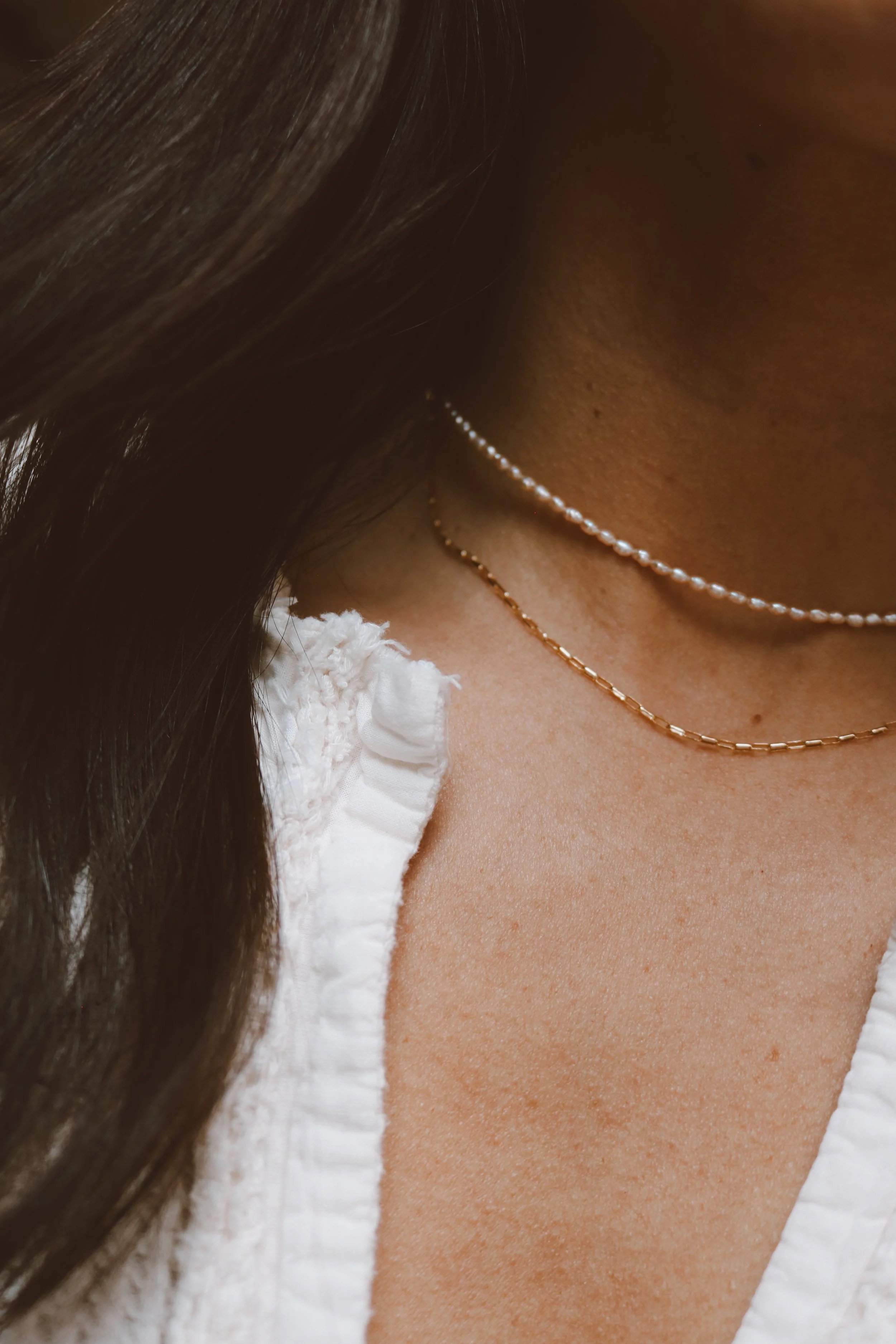 Close-up of a woman's neck with layered necklaces, wearing a white ruffled top and having long dark hair.