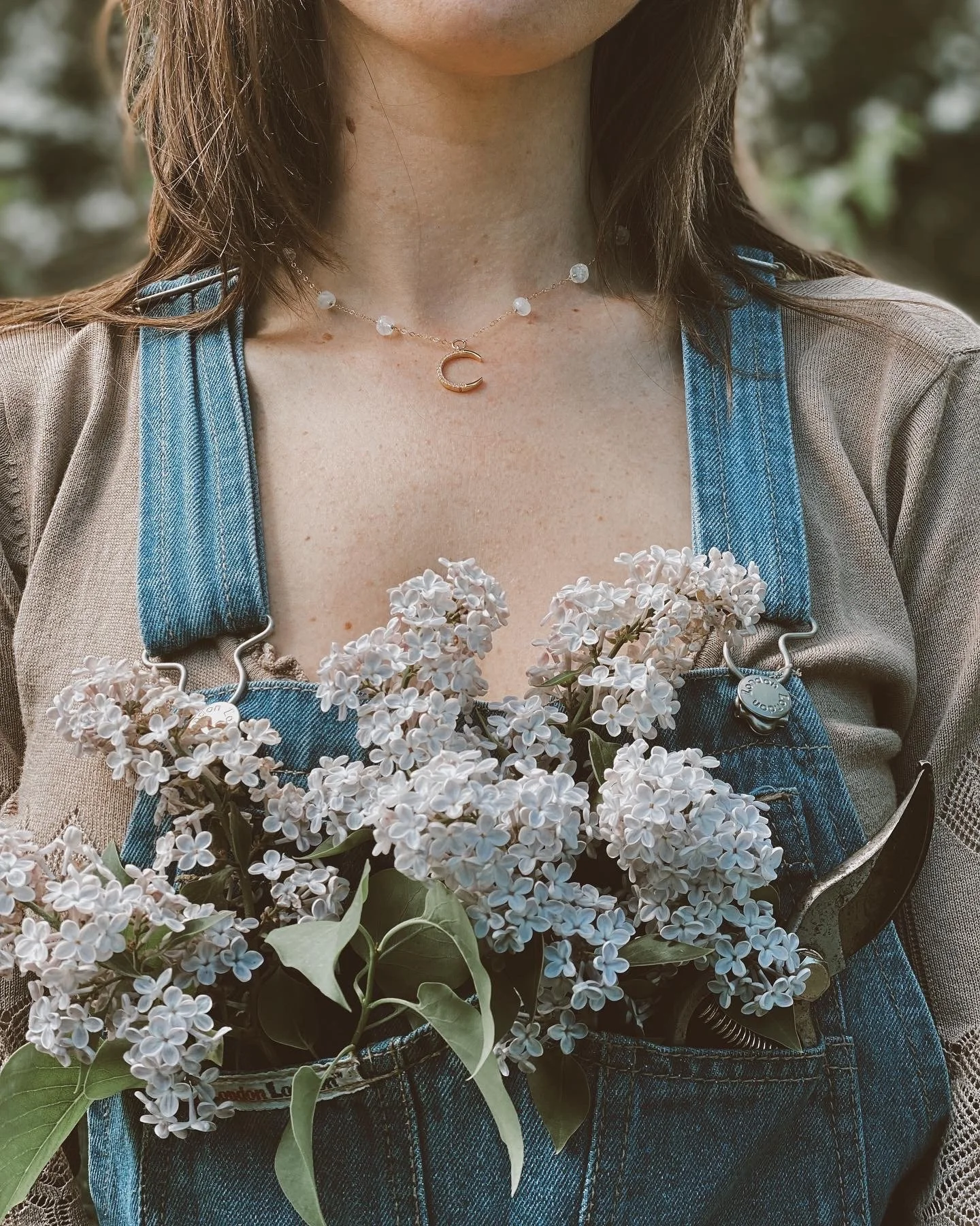 Person wearing a necklace with a crescent moon pendant, holding a bouquet of white lilacs, dressed in denim overalls with a beige top underneath, outdoors.