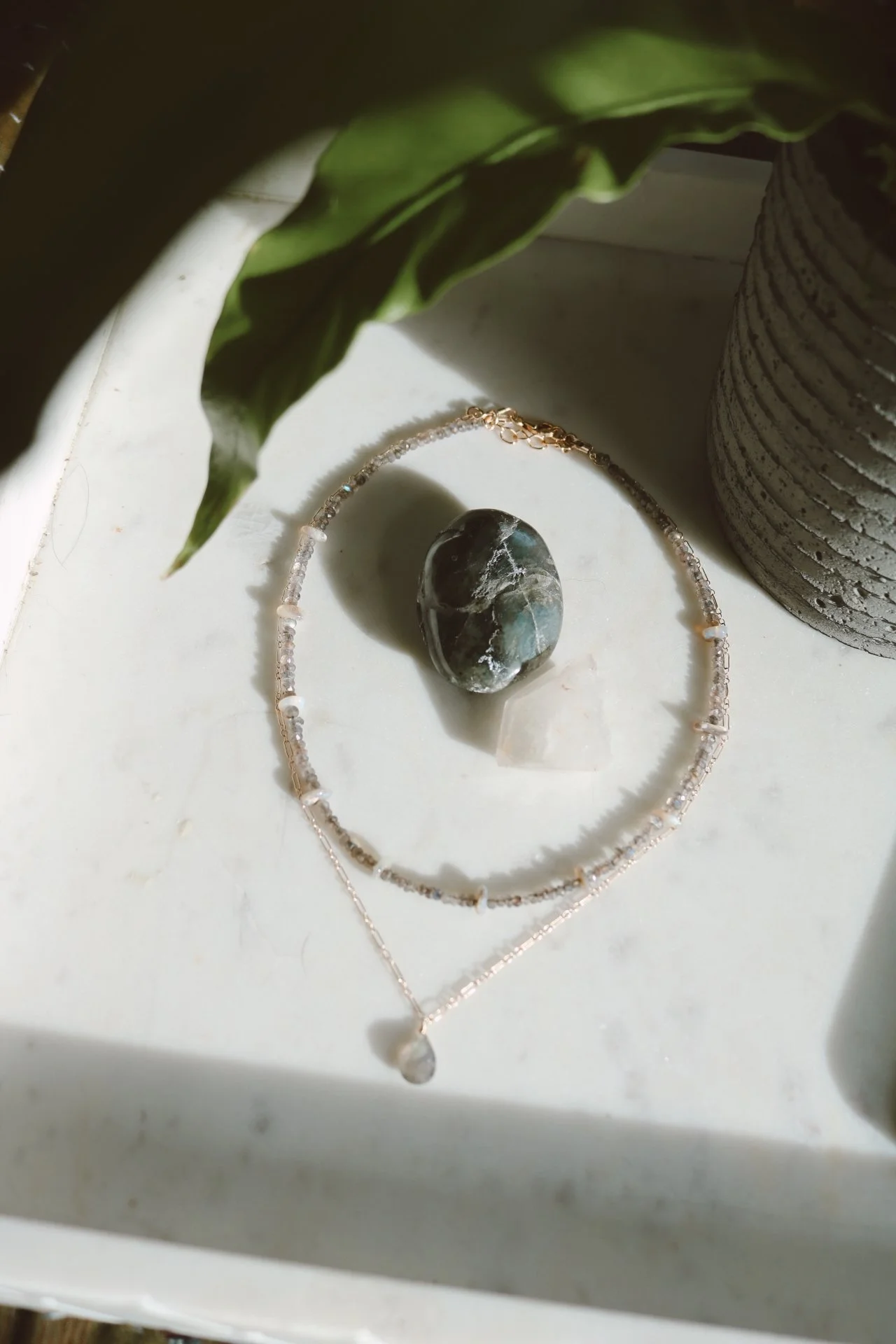 A necklace, a bracelet, and two polished stones on a white surface, with a green plant and a textured gray vase nearby.