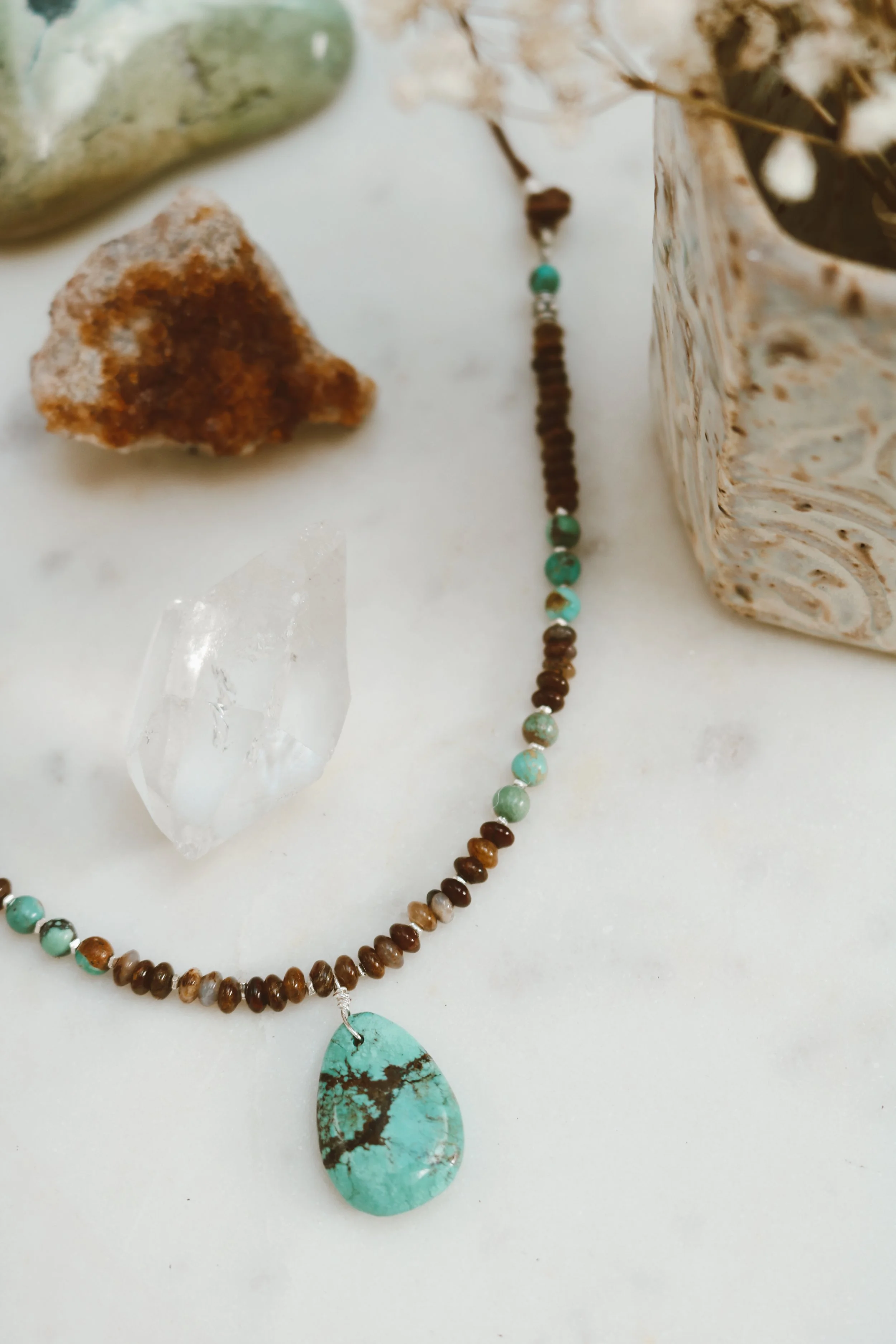 A beaded necklace with a turquoise pendant lying on a white surface, accompanied by a clear quartz crystal, a brown mineral, a green stone, and a ceramic vase with dried flowers.