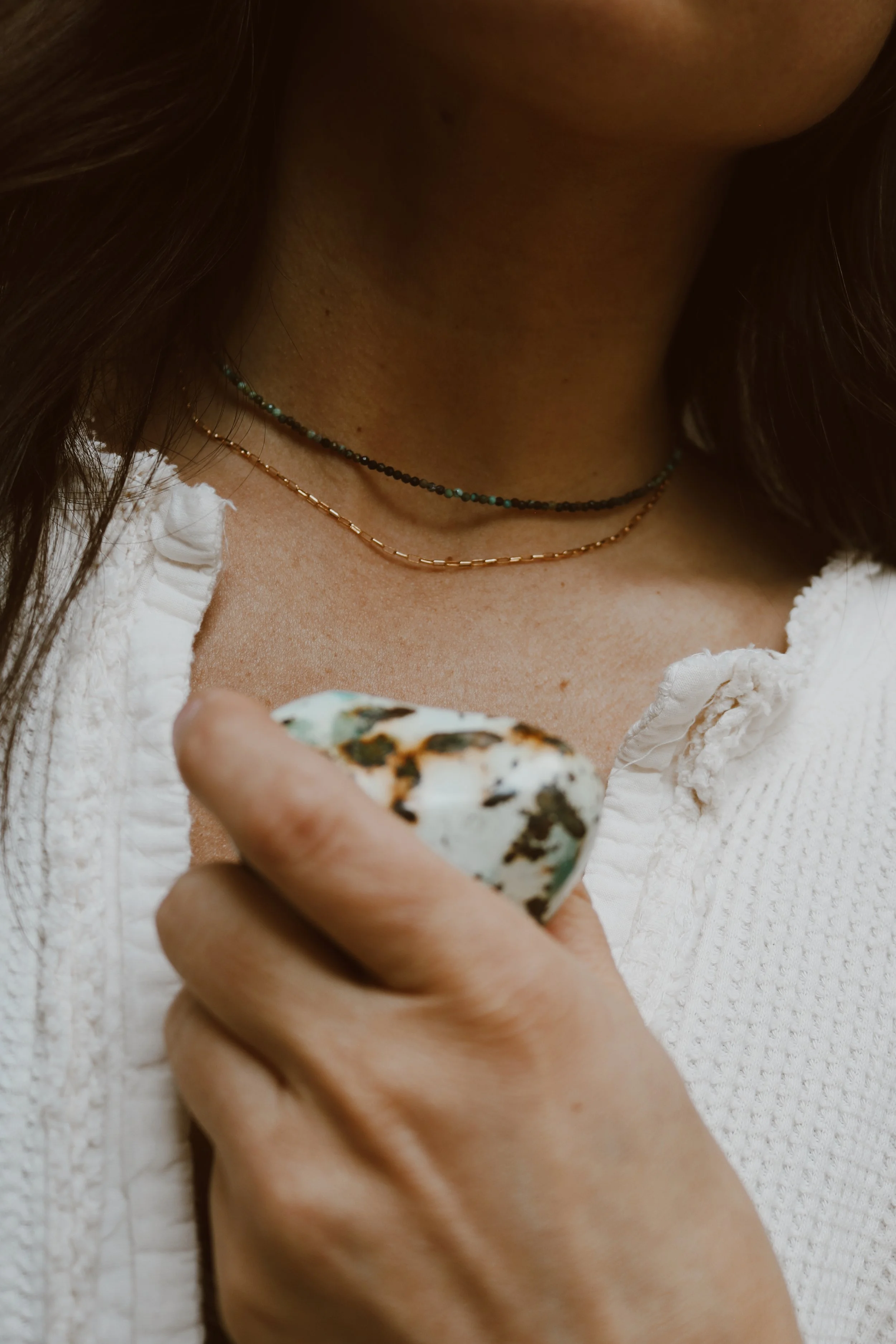 Close-up of a person's neck and hand holding a white and brown patterned stone, wearing layered necklaces and a white textured top.