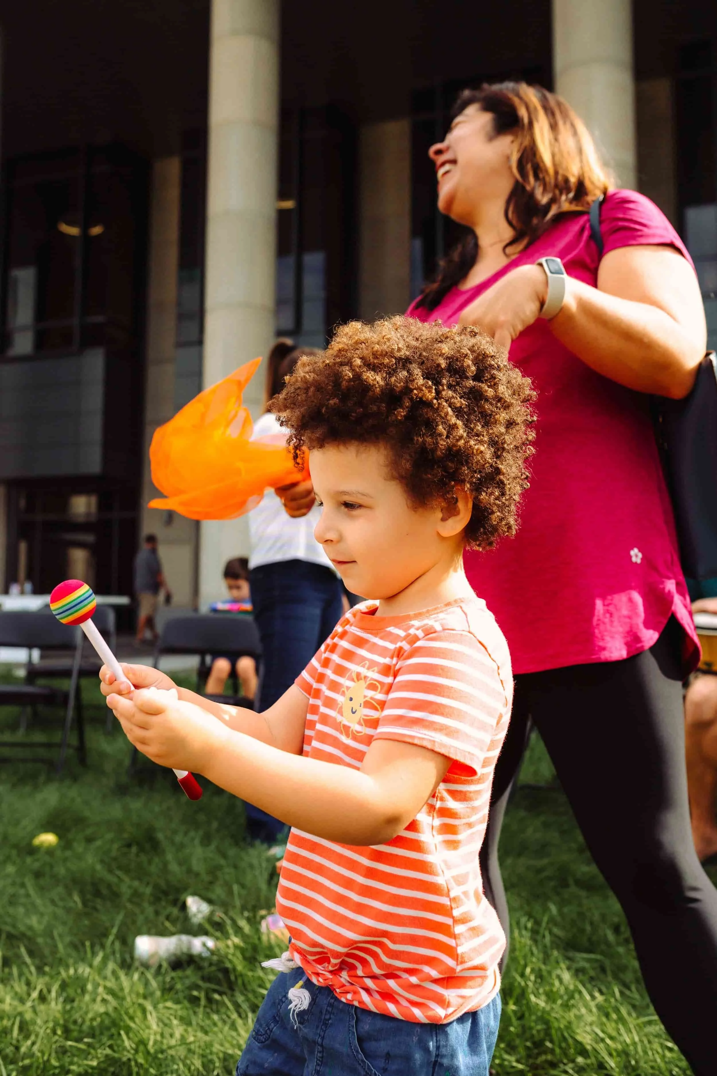 A young boy with curly hair in an orange and white striped shirt looks at a colorful toy while standing on a grassy area, with a woman in a pink shirt and a building in the background.