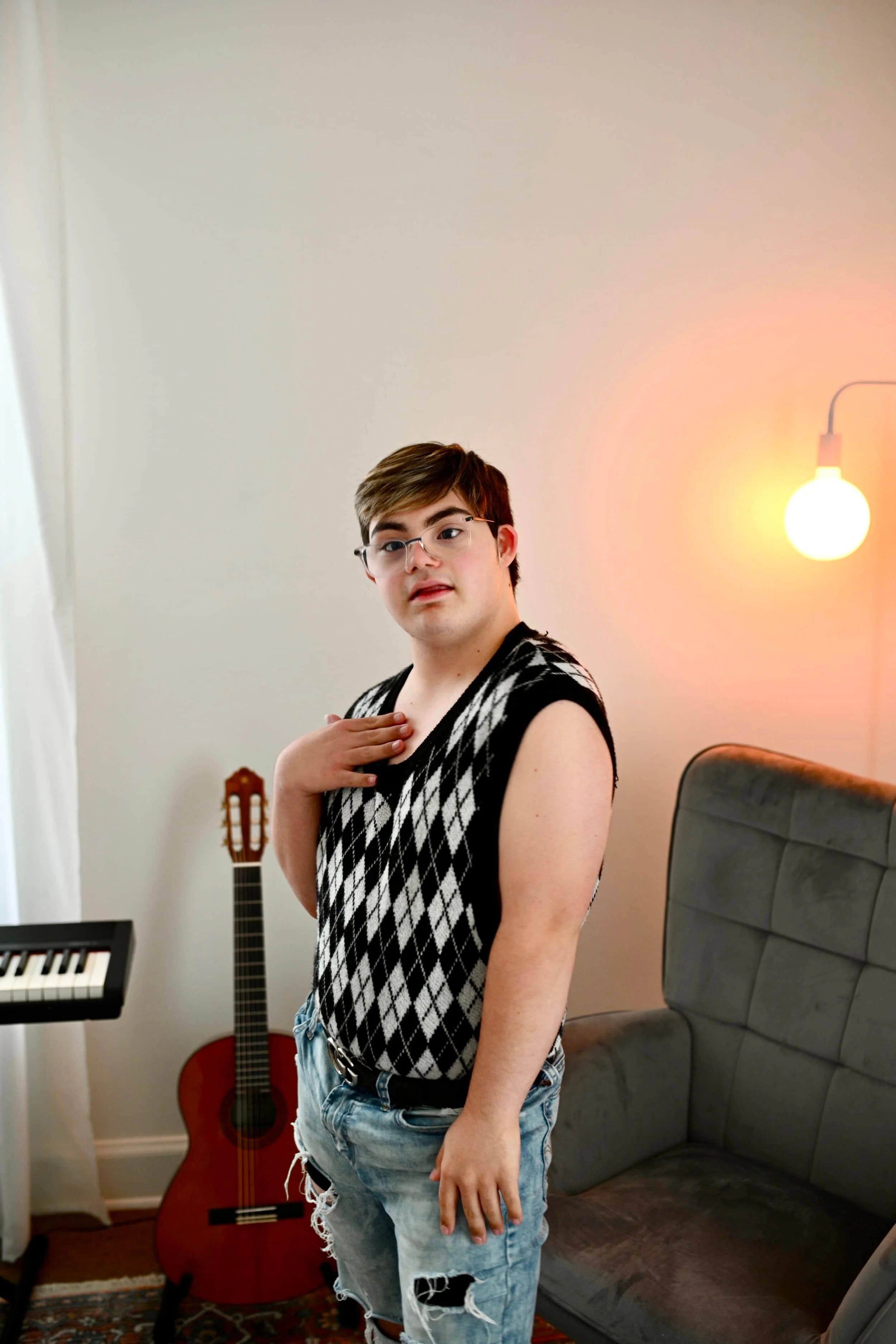 Young man with glasses and brown hair, wearing a sleeveless black and white argyle sweater and ripped denim shorts, indoors near a bed and musical instruments, with a warm glowing lamp in the background.