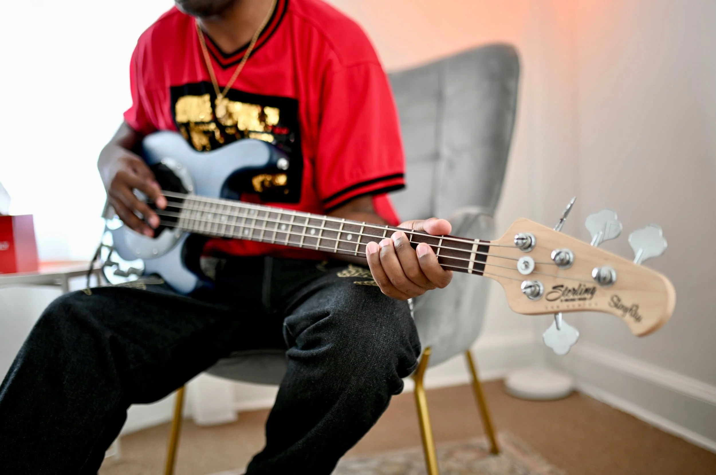 Person in a red shirt plays a silver electric bass on a gray chair indoors, focusing on relaxation and adult mental health Nashville.