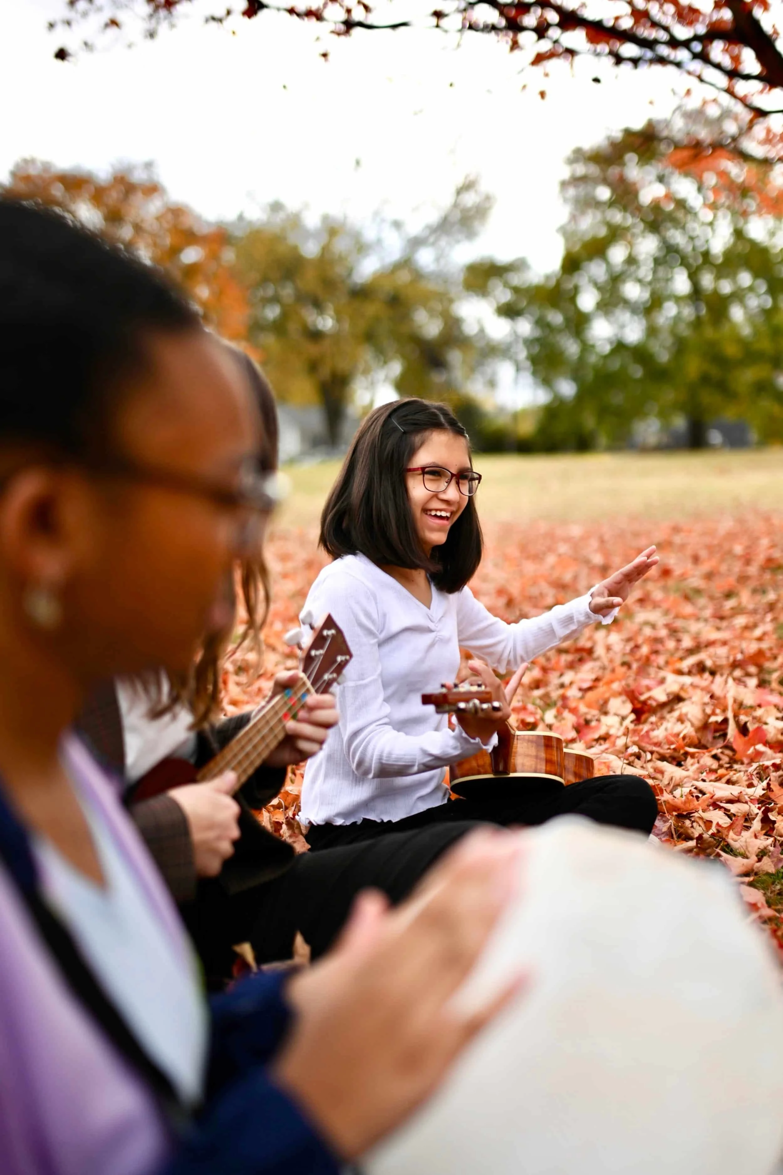 Three women sitting on a bed of autumn leaves outdoors, playing musical instruments and smiling, with a background of trees with fall foliage.