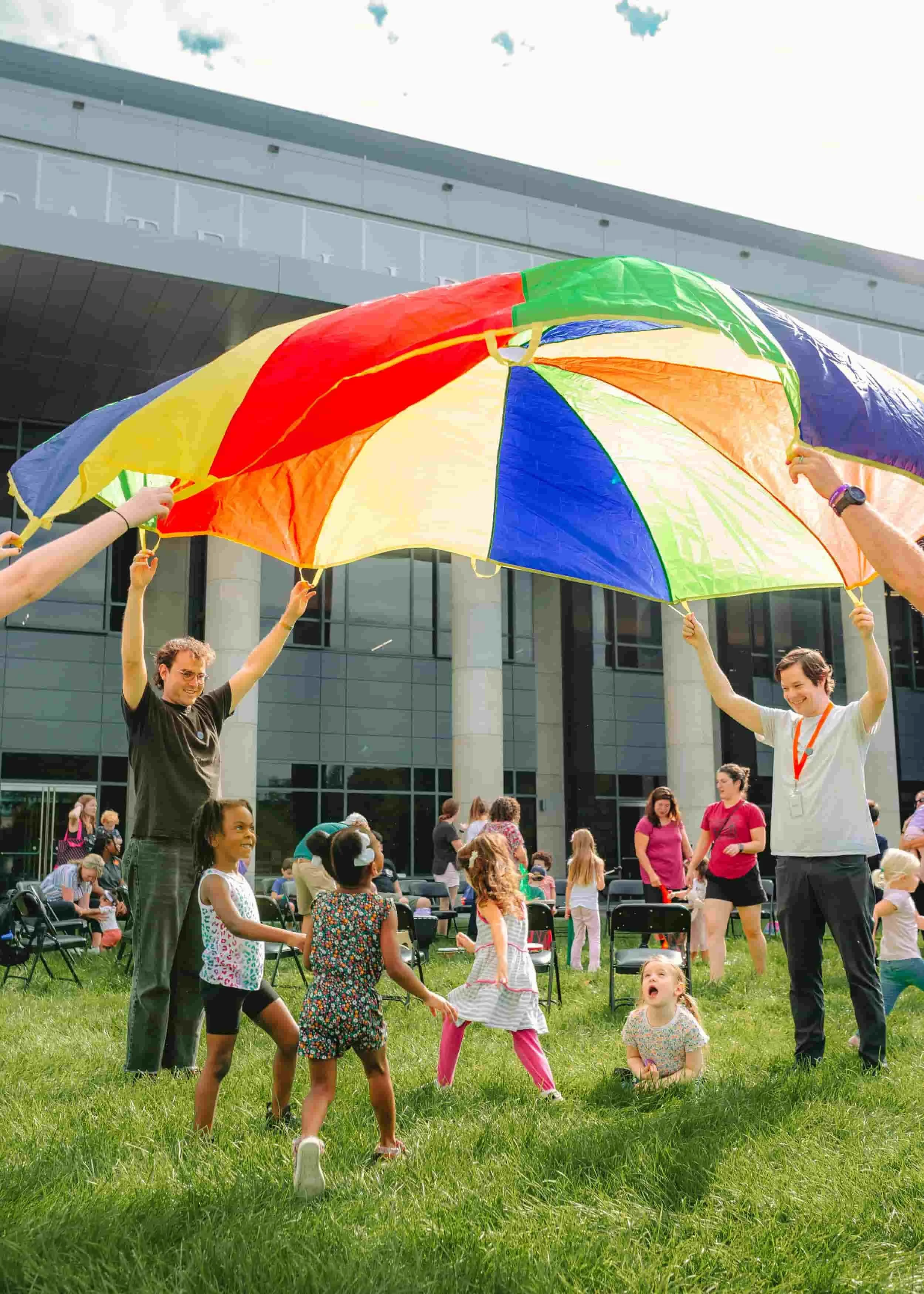 Group of children and adults playing with a large, colorful umbrella outside in a grassy area in front of a modern building.