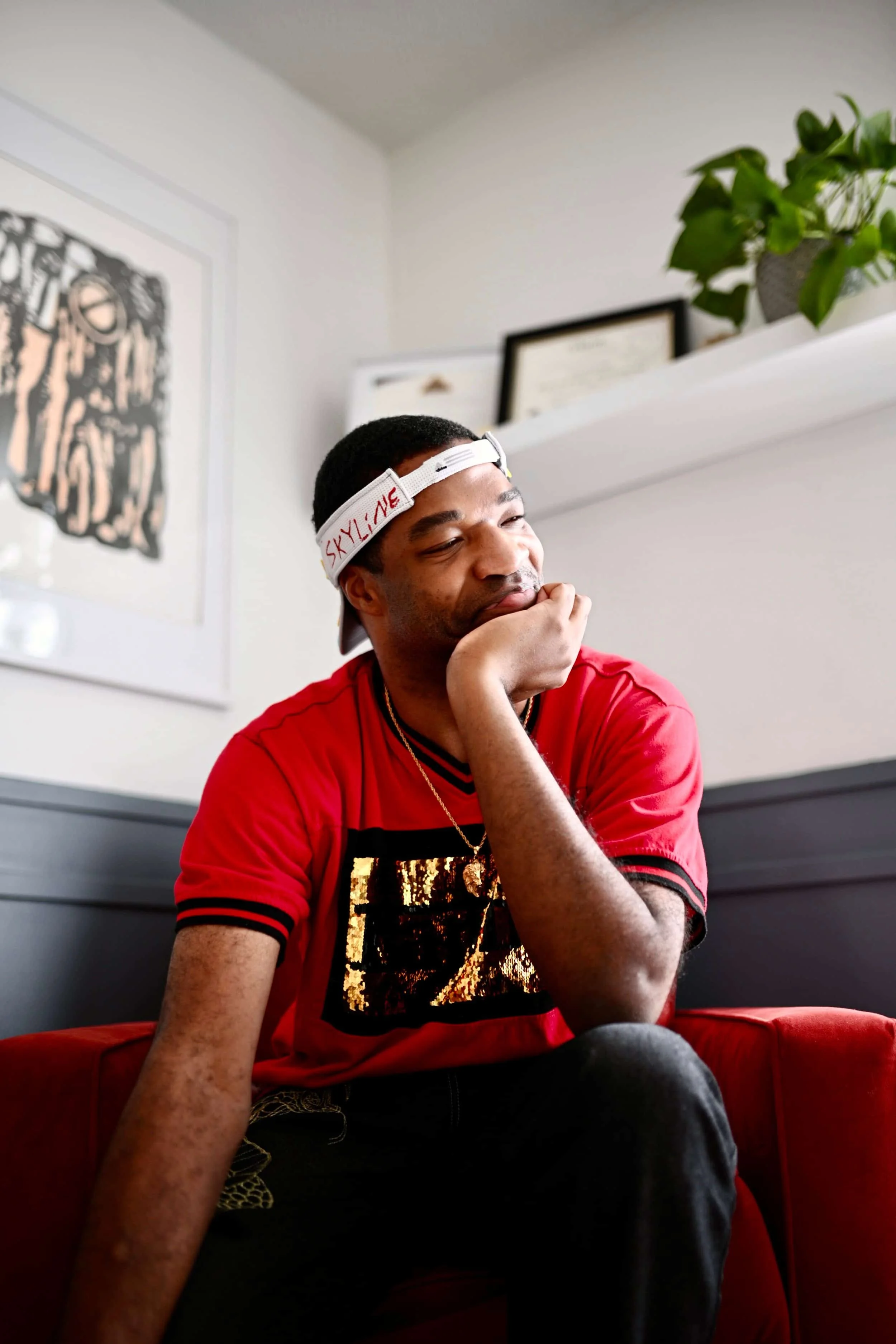 A young man with dark hair wearing a red t-shirt with black sequins and a white visor with the word Skyline stitched in red, sitting on a red chair with his chin resting on his hand, smiling thoughtfully in a room decorated with framed artwork and a potted plant.