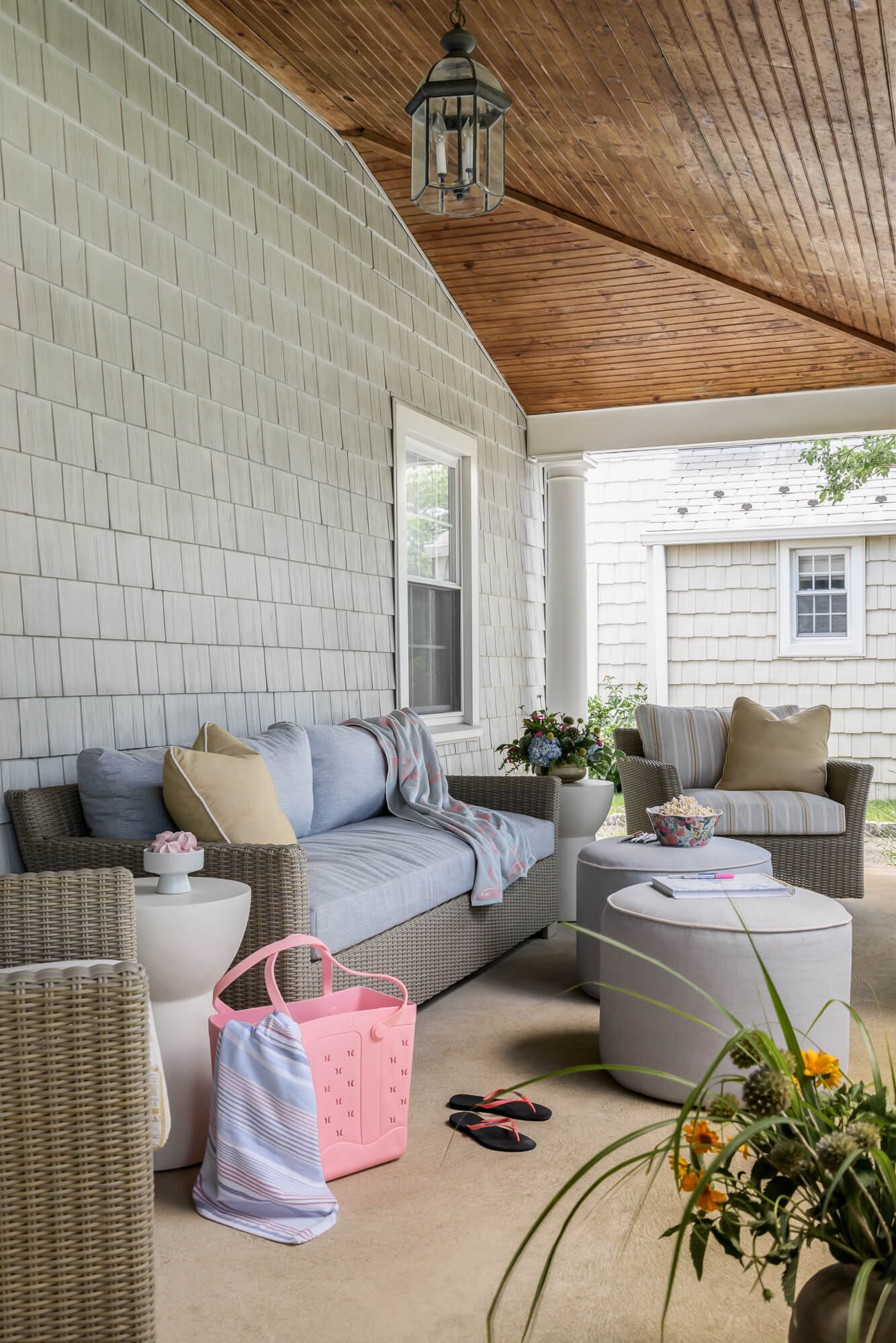An outdoor porch area in Sea Girt, NJ. The porch is designed for a client's beach house in the Jersey by Elements Interior Design Studio and features a wicker outdoor sofa with blue performance fabric and 2 swivel chairs with gray and white striped c