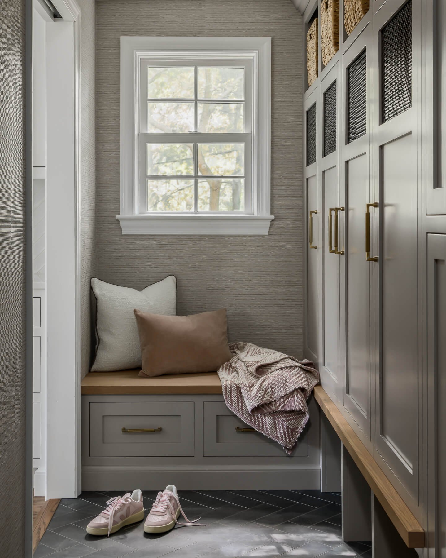 Mudroom renovation in Cranford, NJ by Elements Interior Design Studio. There are greige cubbies with mesh inserts and brass hardware and black herringbone tile. The photo looks towards a window seat with drawers below and 2 pillows and a blanket.