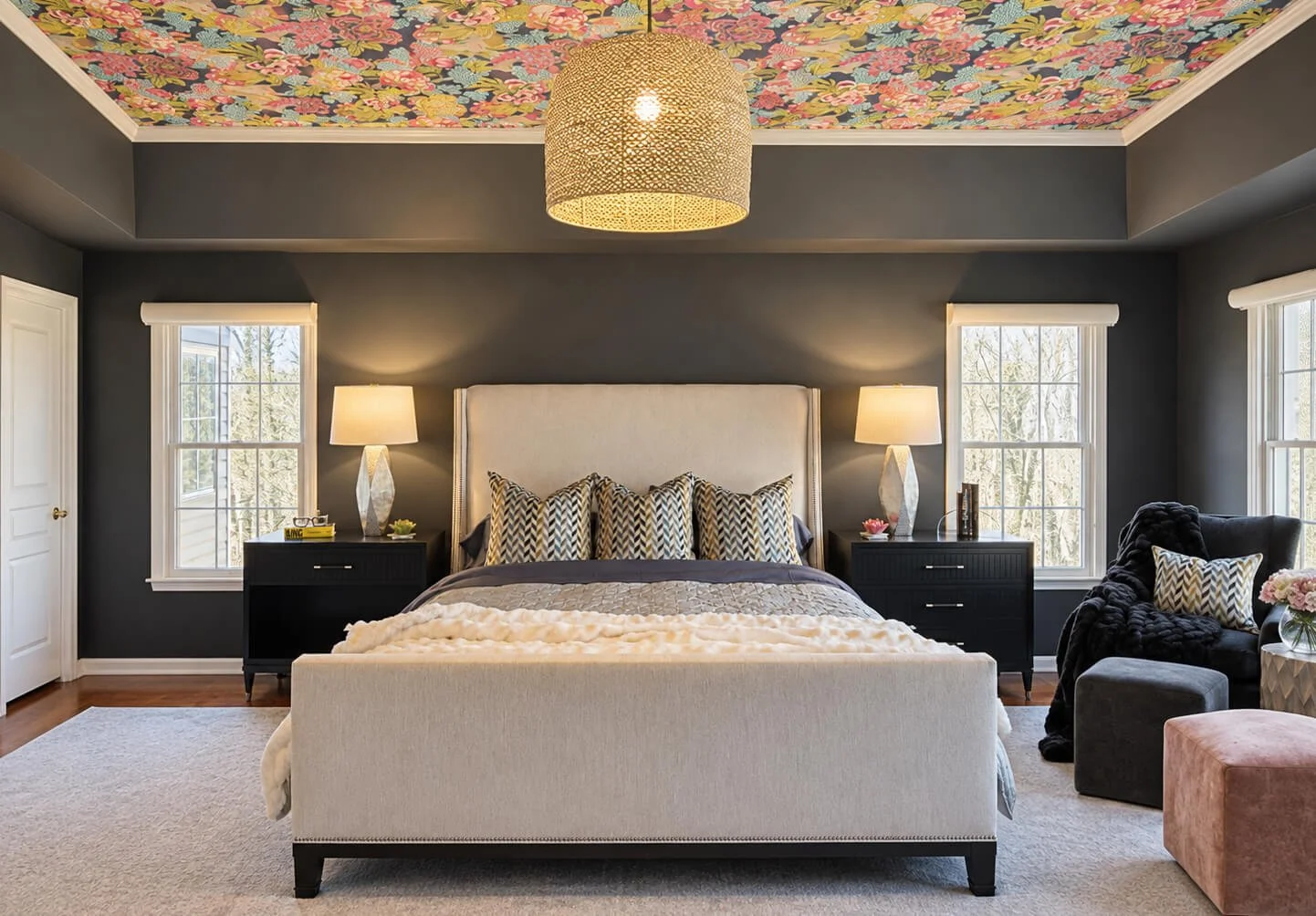 Primary bedroom design project in Warren, NJ by Elements Interior Design Studio. The ceiling has a colorful floral wallpaper and the walls are painted a dark charcoal gray. There is a large natural chandelier pendant over the bed. The bed has a linen