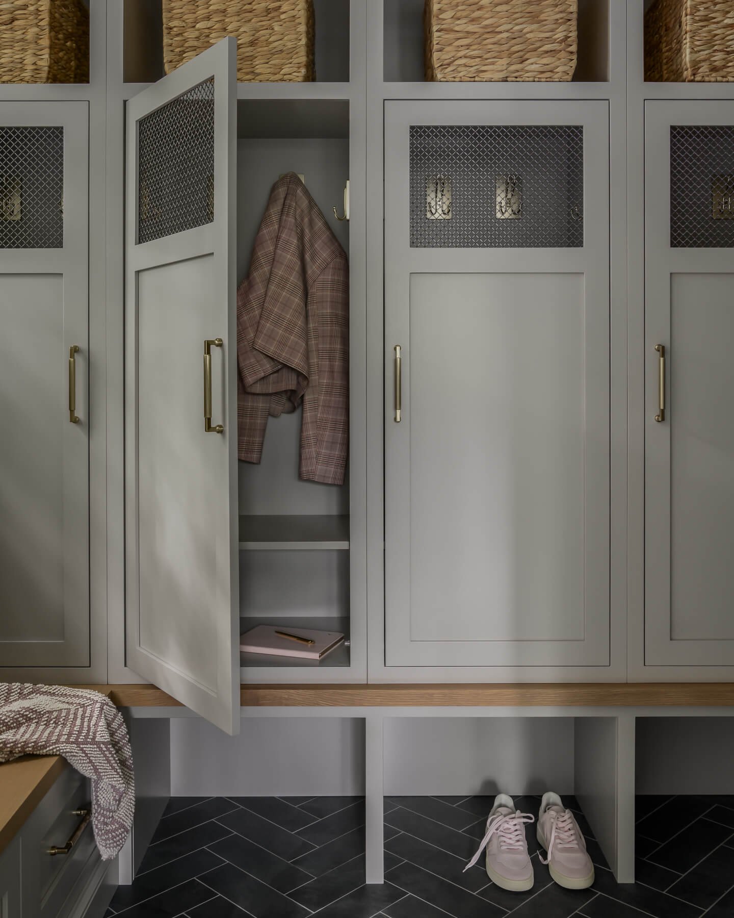 Mudroom renovation in Cranford, NJ by Elements Interior Design Studio with greige cubbies with brass hardward and mesh inserts and a black tile in a herringbone pattern. One of the doors is open showing a pink jacket hanging inside.