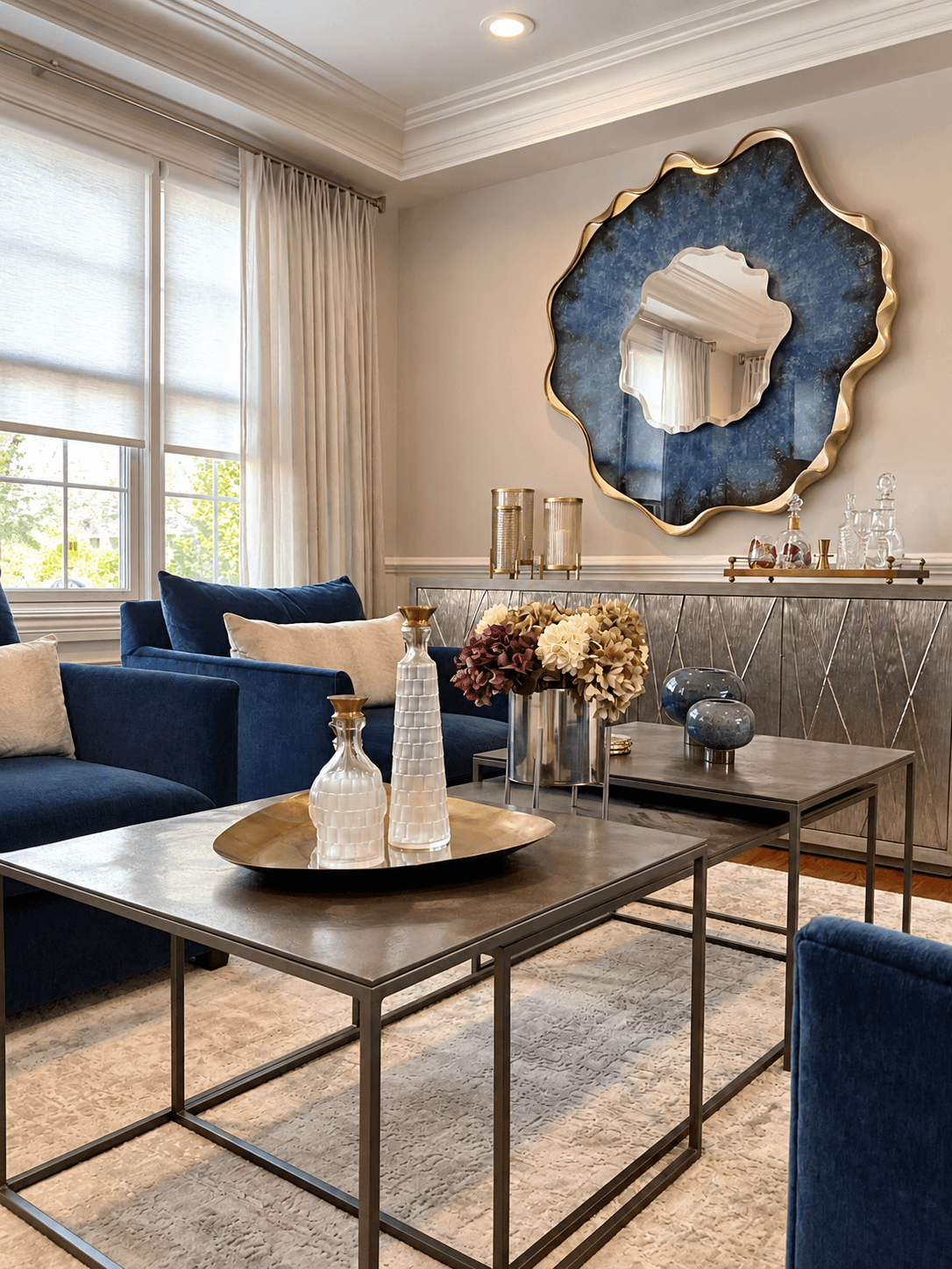 Living room design project in Scotch Plains, NJ by Elements Interior Design Studio featuring a modern statement blue and gold mirror on the wall above a metallic console table. The table has decor objects on top. There are 2 nesting brass coffee tabl