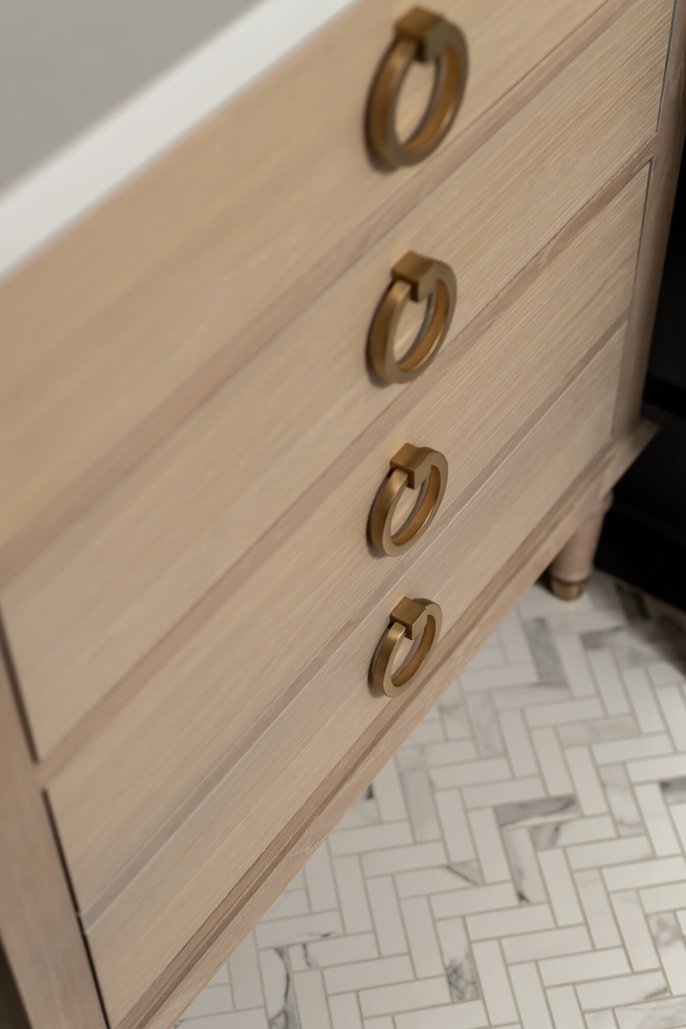Close-up of birch wood vanity with brass ring pulls and a white speckled herringhone tile from a powder room renovation in Cranford, NJ by Elements Interior Design Studio.