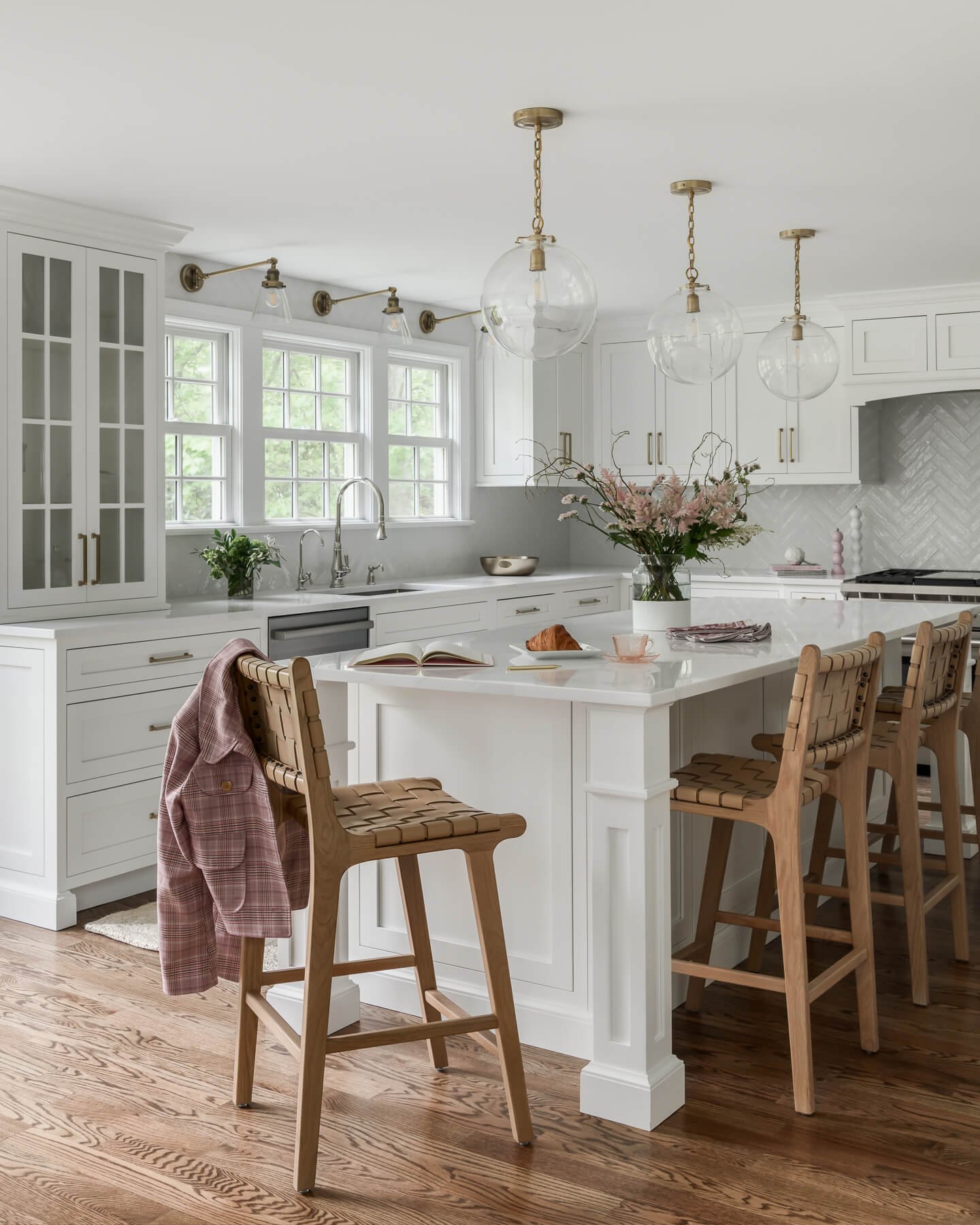 Kitchen renovation in Cranford, NJ by Elements Interior Design Studio. The cabinets and countertops are white with a white glossy herringbone backsplash. There are woven leather counter stools around the island 3 glass globe pendant lights above the 