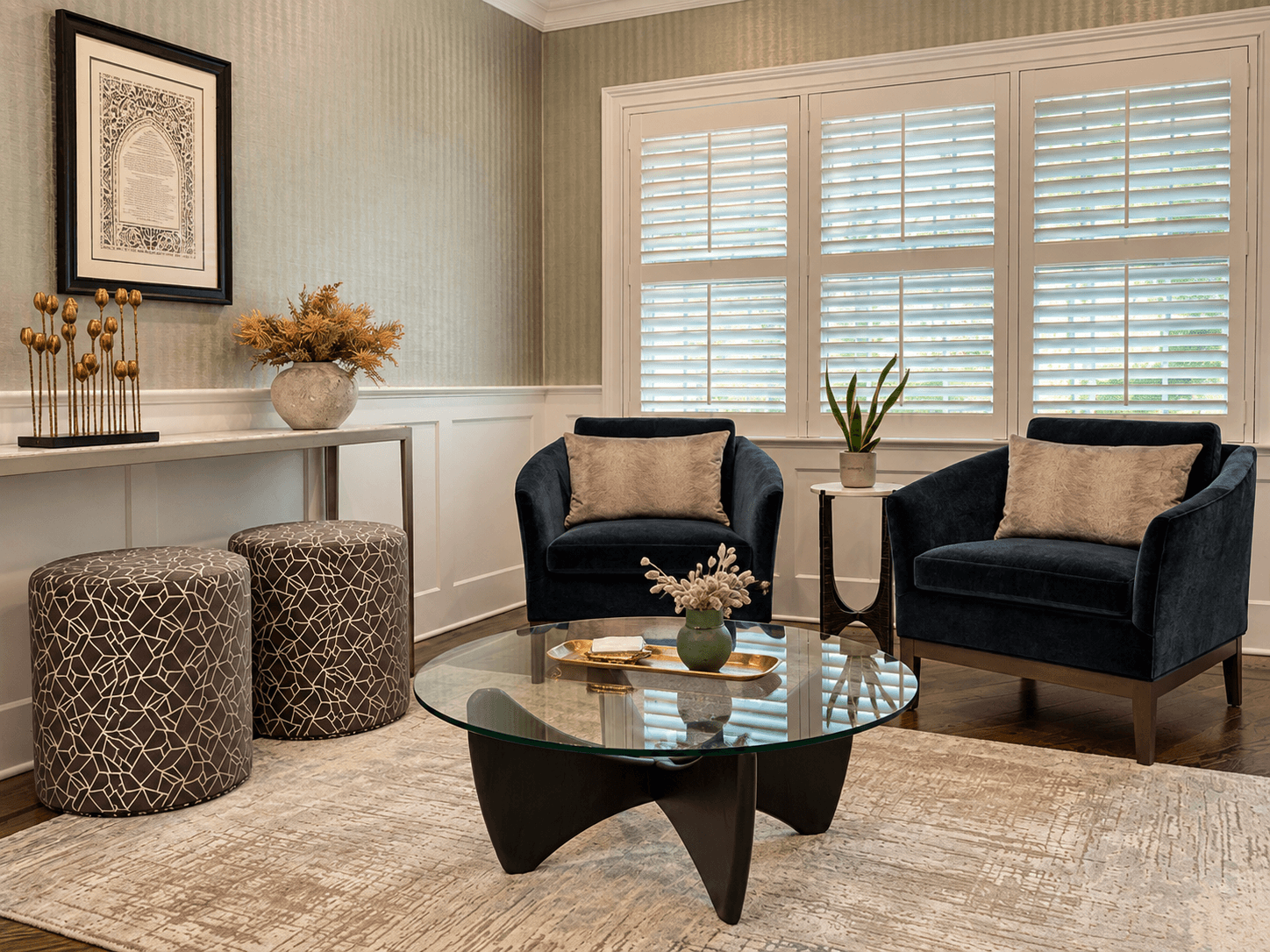 Modern living room interior design project in Westfield, NJ by Elements Interior Design Studio. There are 2 dark blue club chairs with mauve throw pillows in front of a large window with shutters. There is a round, glass coffee table in front of the 