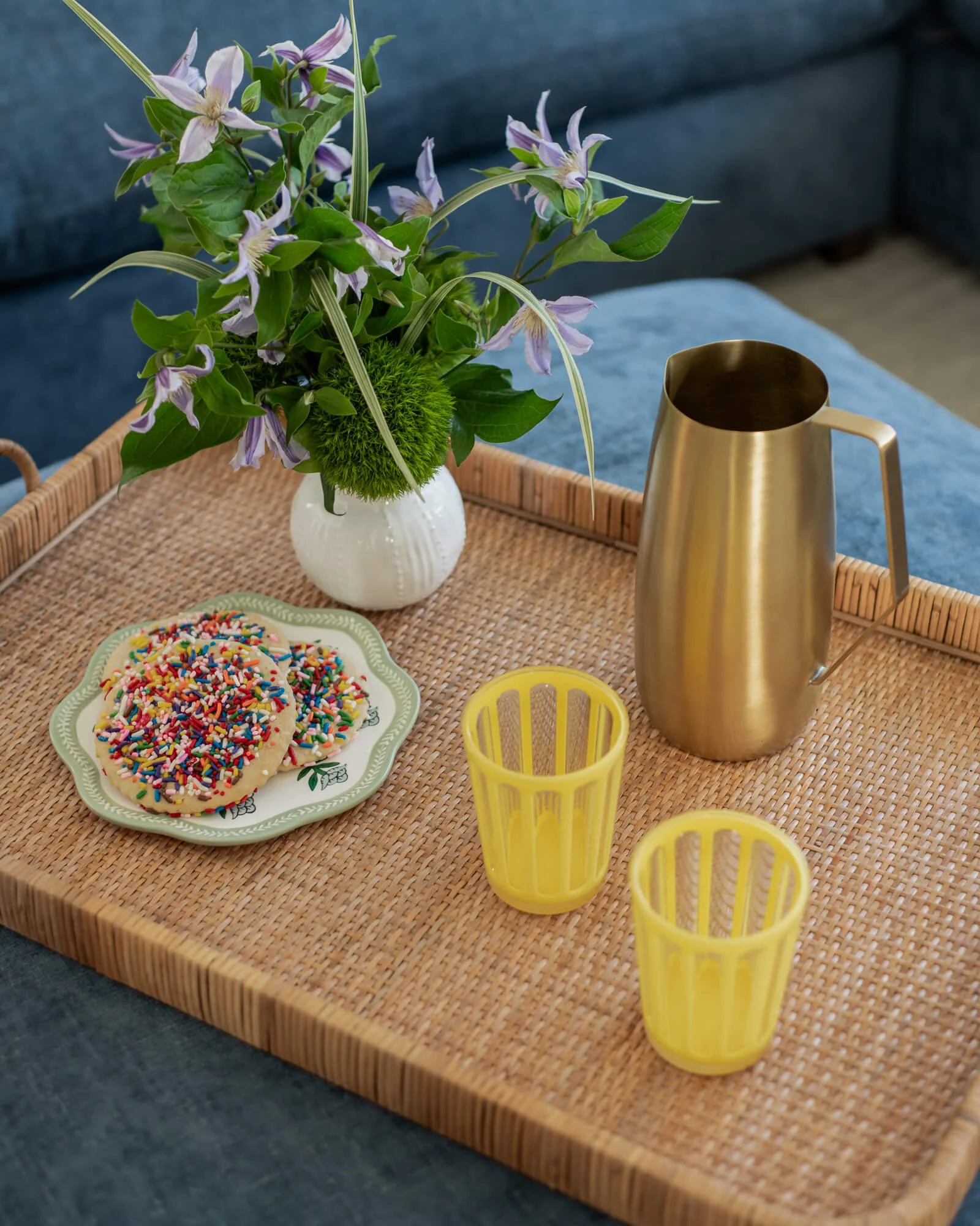 A close up of a dark blue ottoman from a coastal family room interior design project in the Jersey Shore by Elements Interior Design Studio. There is wicker tray with a white vase holding purple and green flowers, two yellow cups, a gold metal pitche