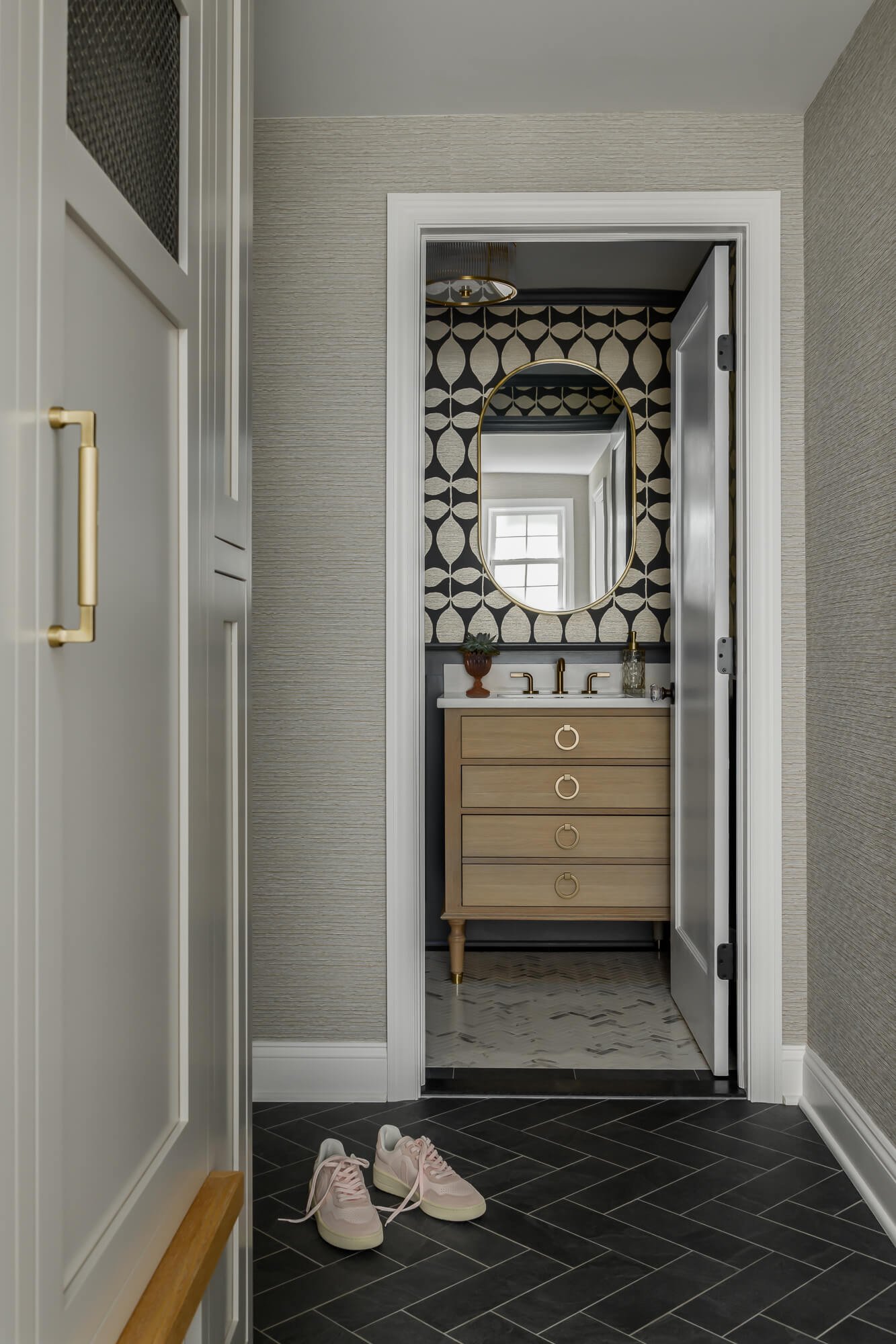 Home renovation in Cranford, NJ by Elements Interior Design Studio. Photo shows a hallway with white cabinetry and black tile floor looking into a powder room with a bold black and beige patterned wallpaper, a birch vanity and an brass oval mirror.