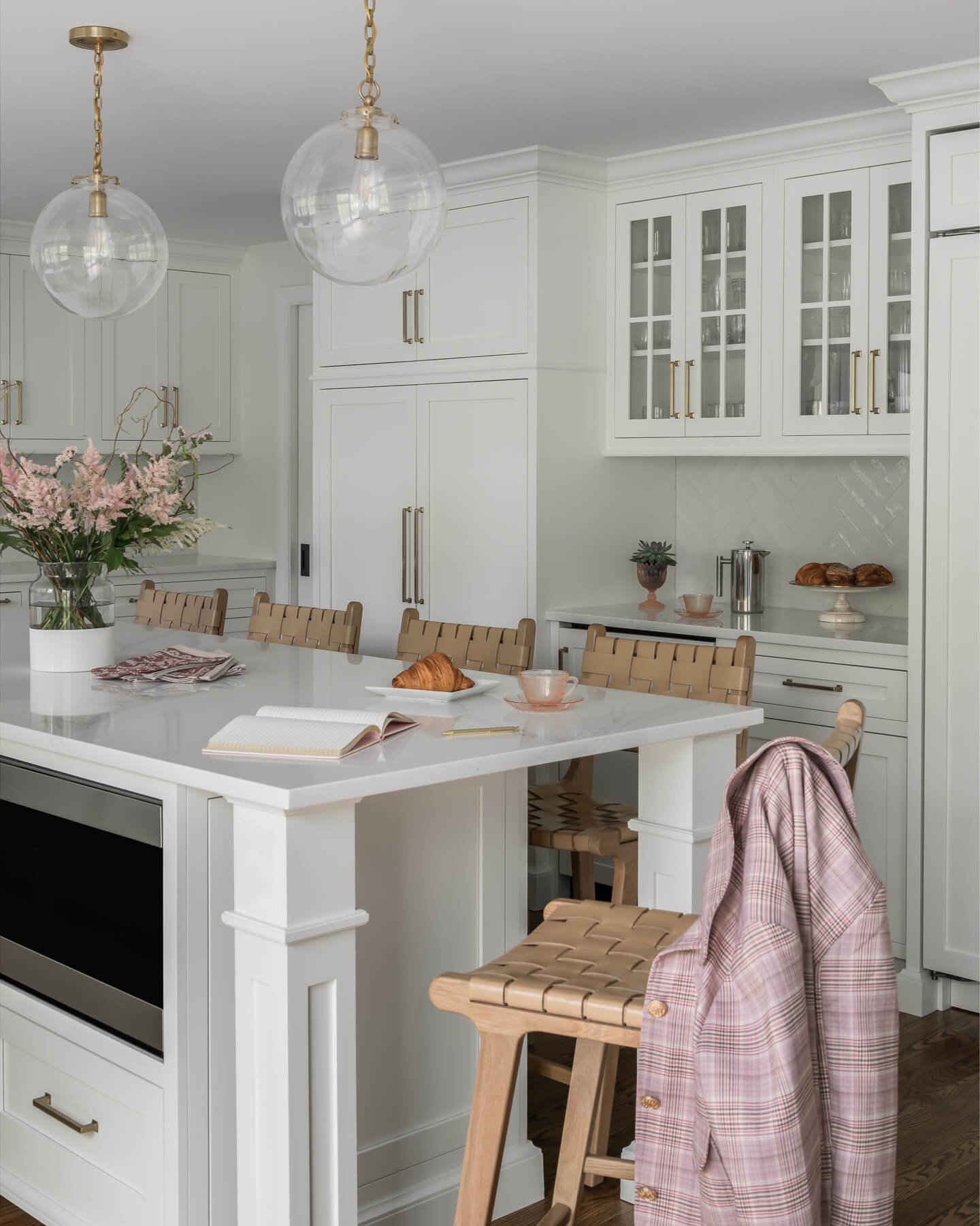 Kitchen renovation in Cranford, NJ by Elements Interior Design Studio. The cabinets and countertops are white with a white glossy herringbone backsplash. There are woven leather counter stools around the island, with a pink jacket on one of the stool