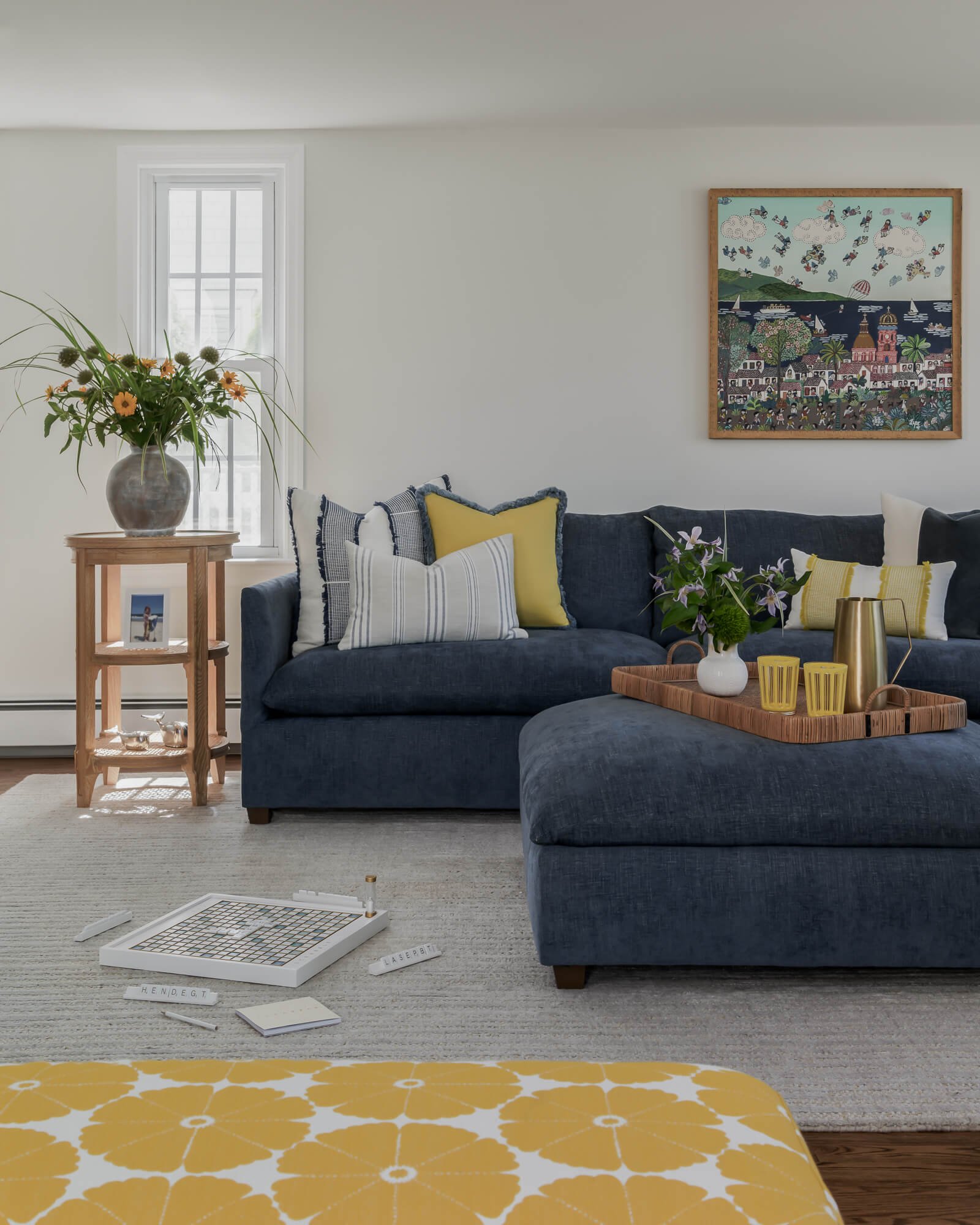 
Coastal style family room interior design project for a client's beach house in Sea Girt, NJ by Elements Interior Design Studio with large windows, dark blue velvet sectional sofa with yellow and blue throw pillows, and ottoman with a rattan tray wi
