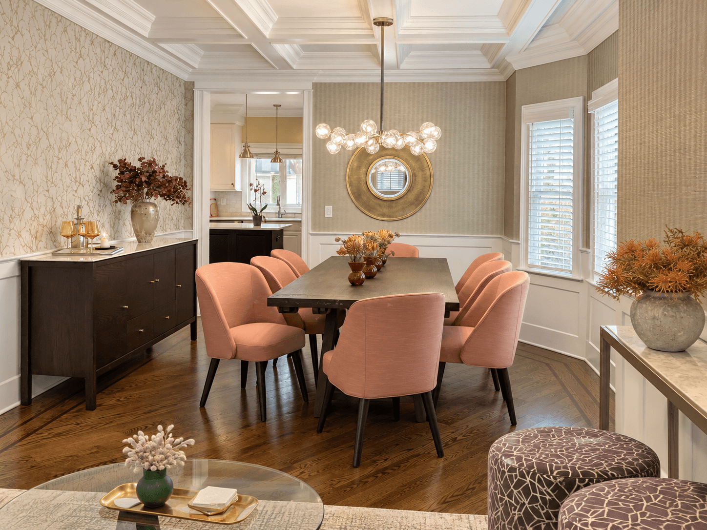 Dining room design project in Westfield, NJ, by Elements Interior Design Studio. There is a coffered ceiling and a long brown wood table with modern chairs with coral upholstery. The walls are wallapered in a subtle metallic print. There is a dark br