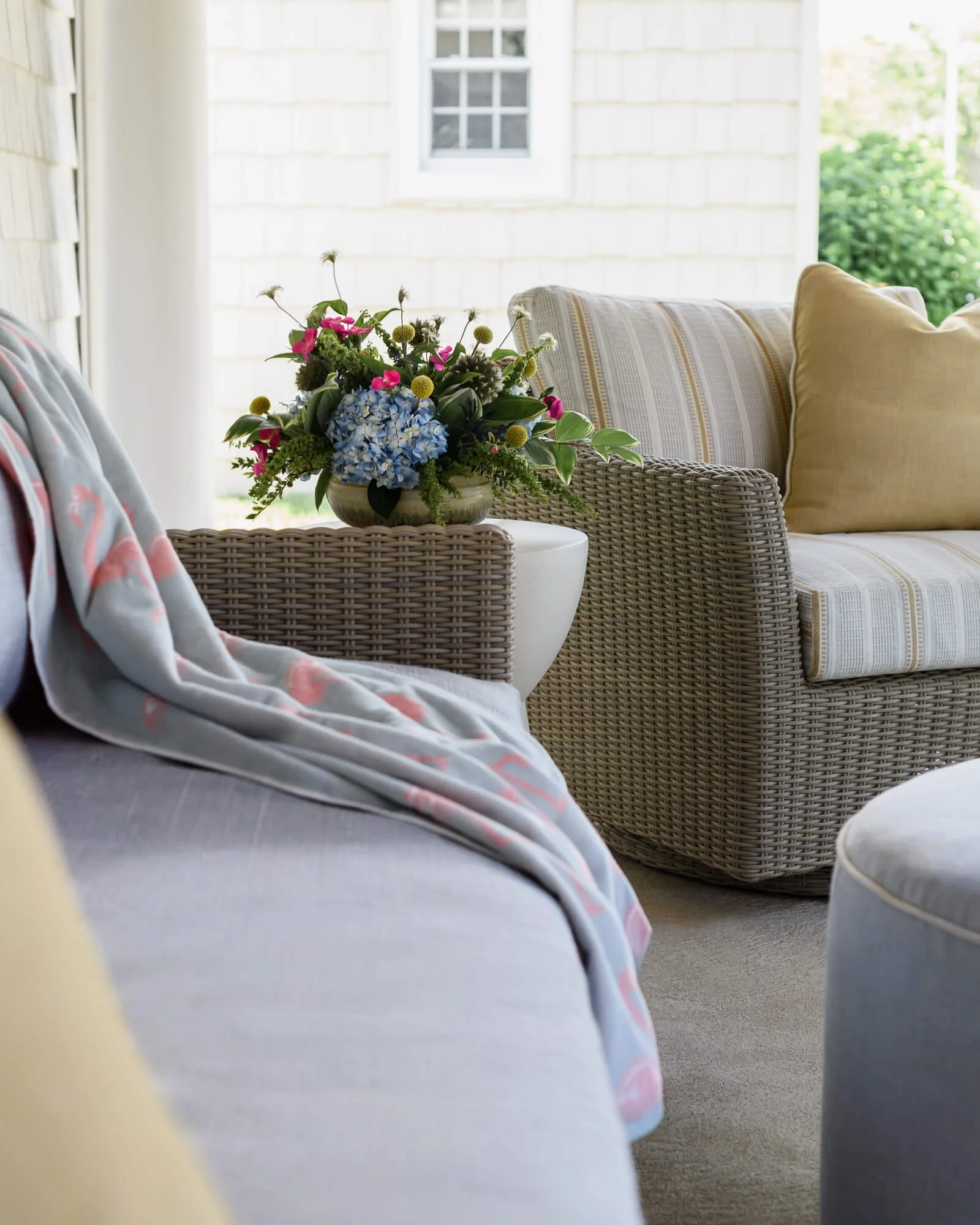 View of a beach home porch design project in Sea Girt, NJ by Elements Interior Design Studio. There is a wicker chair with yellow and gray striped cushion with a yellow throw pillow, and a side table with a vase of colorful flowers. The porch has a w