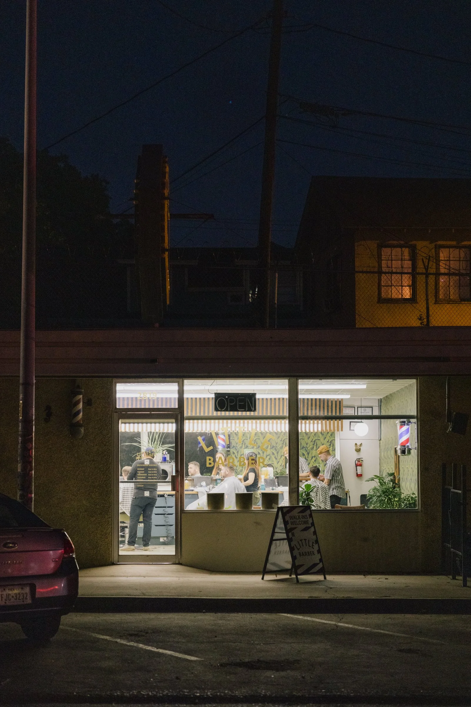 Night exterior of Little Barber barbershop in Austin Texas with warm interior lighting visible through the windows.