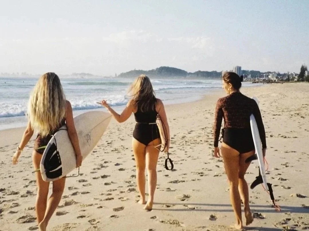 Three women walking along the beach with surfboards, dressed in swimsuits and beachwear, with the ocean and a distant hillside in the background.