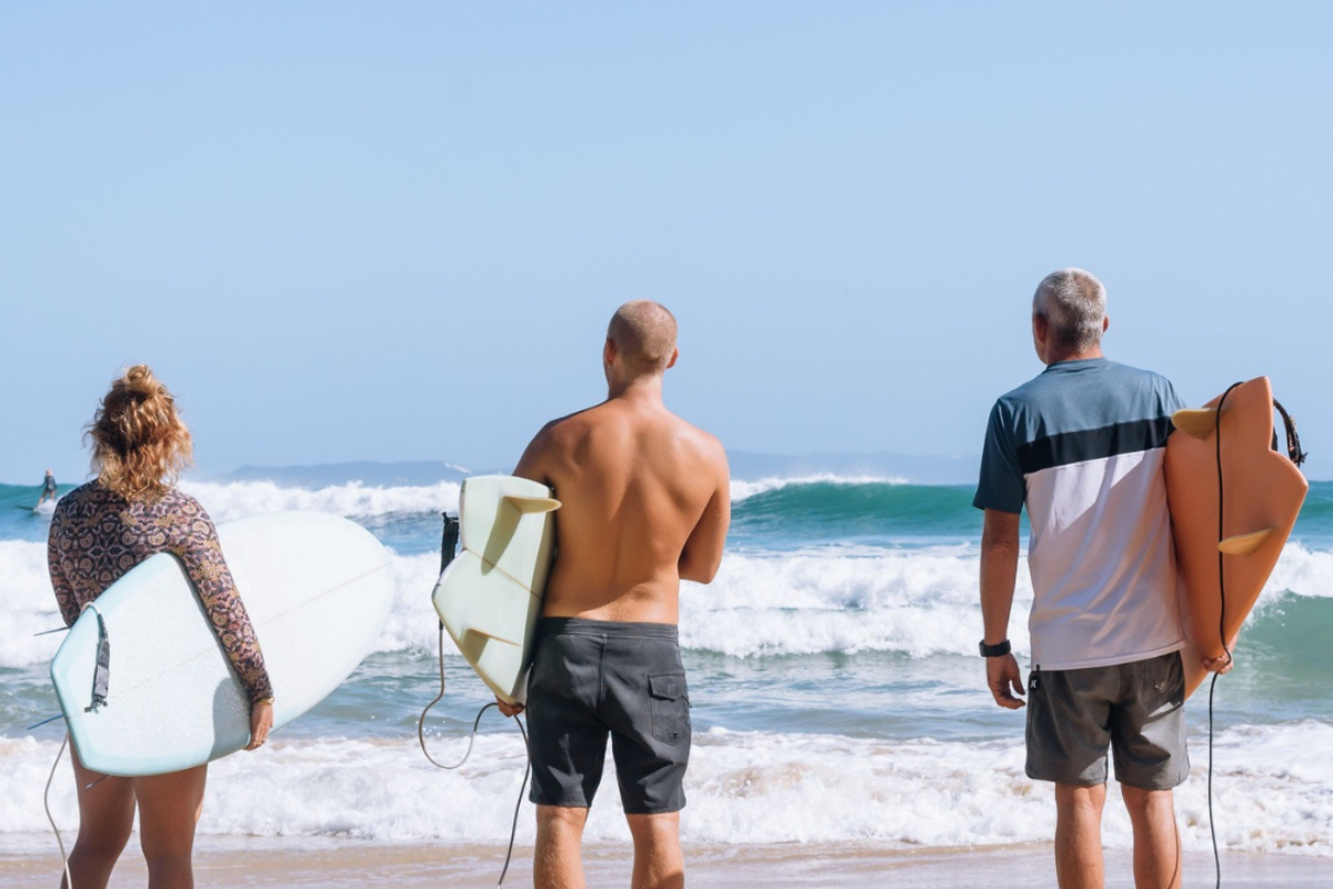 Three surfers walking into the ocean with surfboards on a sandy beach.