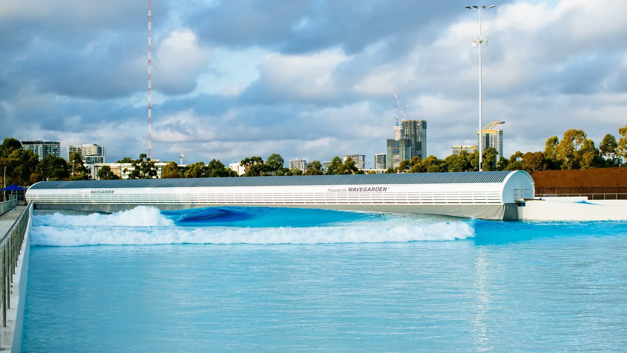 A large Wavegarden wave pool with a city skyline in the background, including high-rise buildings under a cloudy sky.