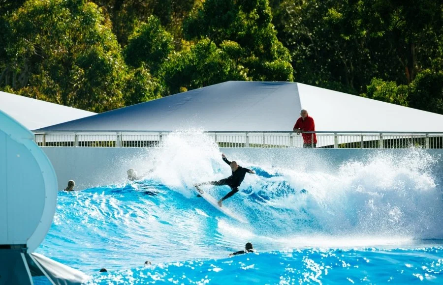 A person is surfing a wave at a water park, with a lifeguard or staff member watching from a platform nearby. Trees are visible in the background.