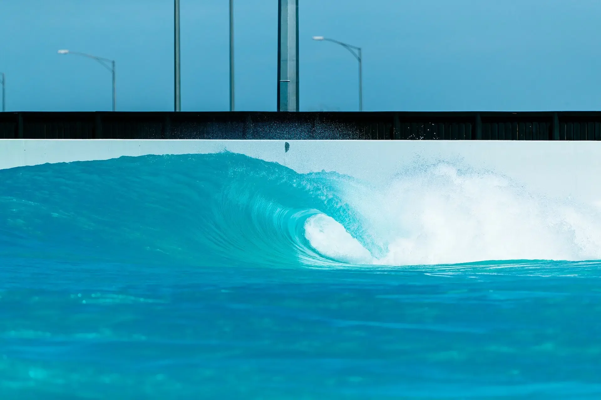 A large ocean wave curling in turquoise water, with a billboard or large screen displaying a plain blue sky with some streetlights in the background.