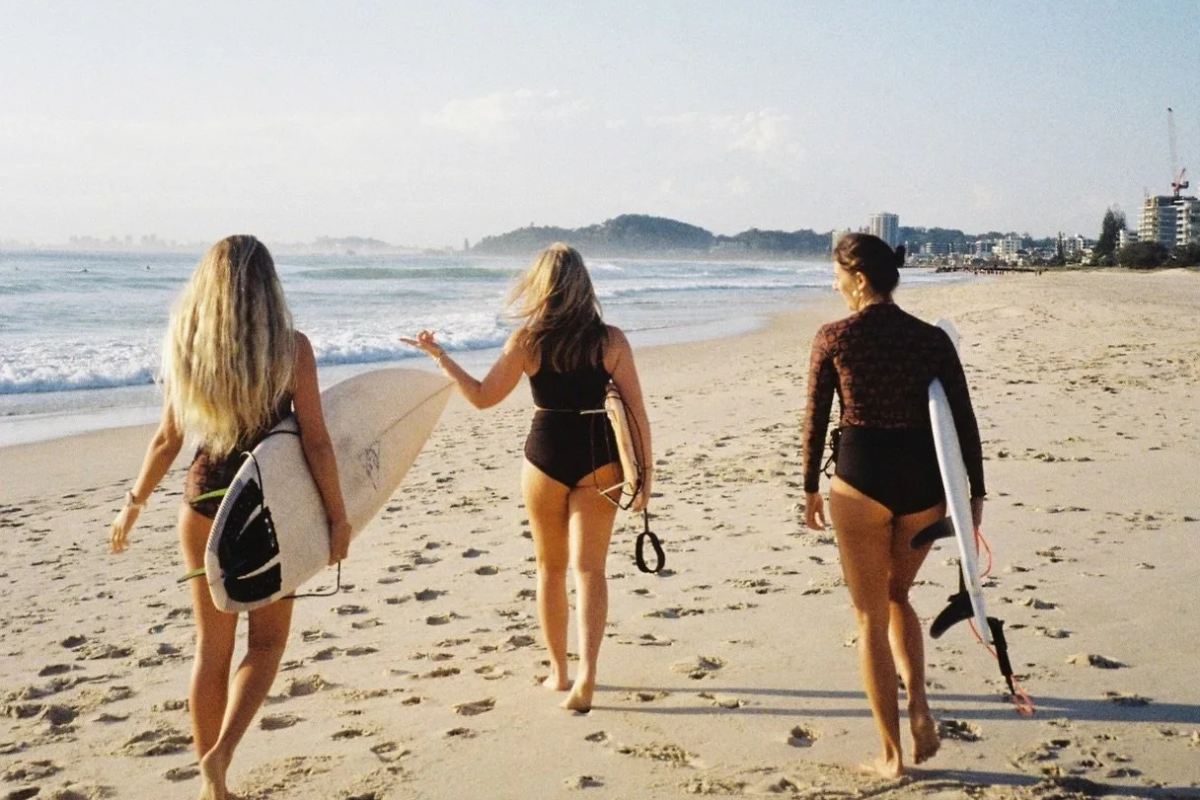 Three women walking on the beach carrying surfboards, one women leading the others, with buildings and a hill in the background.