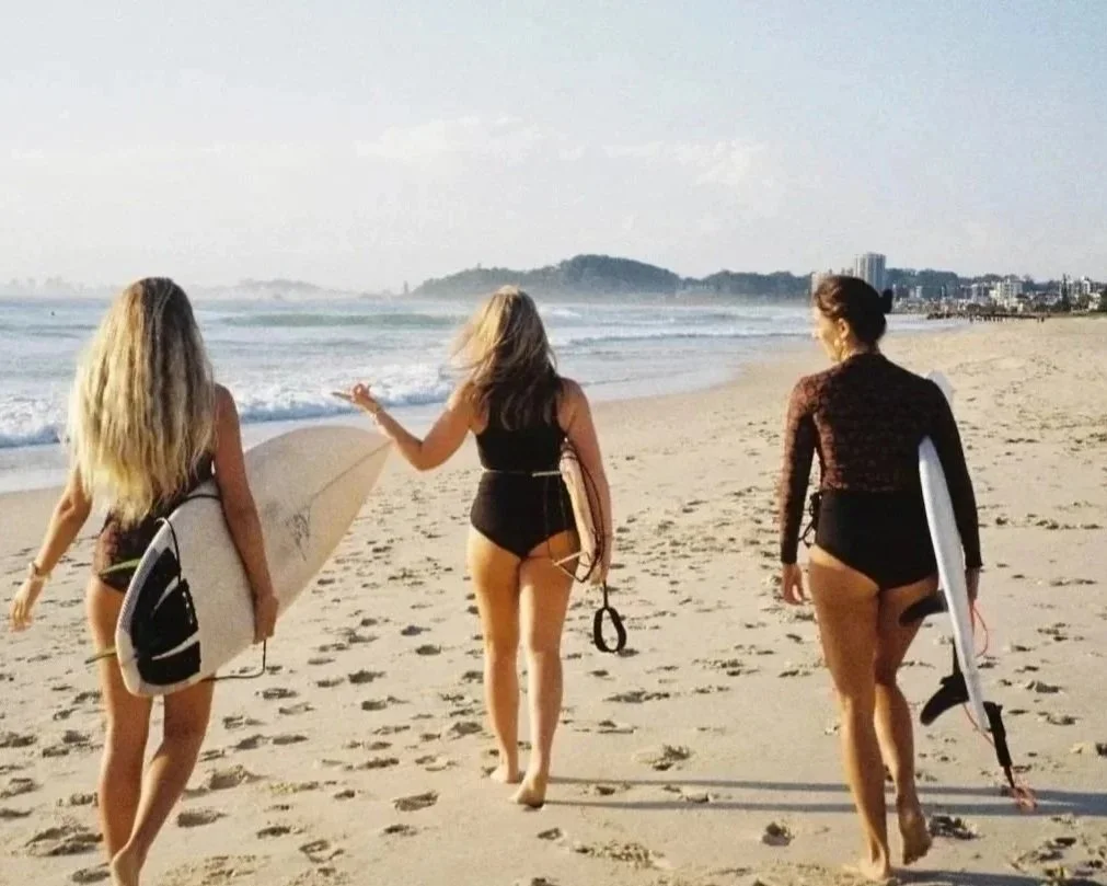 Three women walking along the beach with surfboards, dressed in swimsuits and beachwear, with the ocean and a distant hillside in the background.