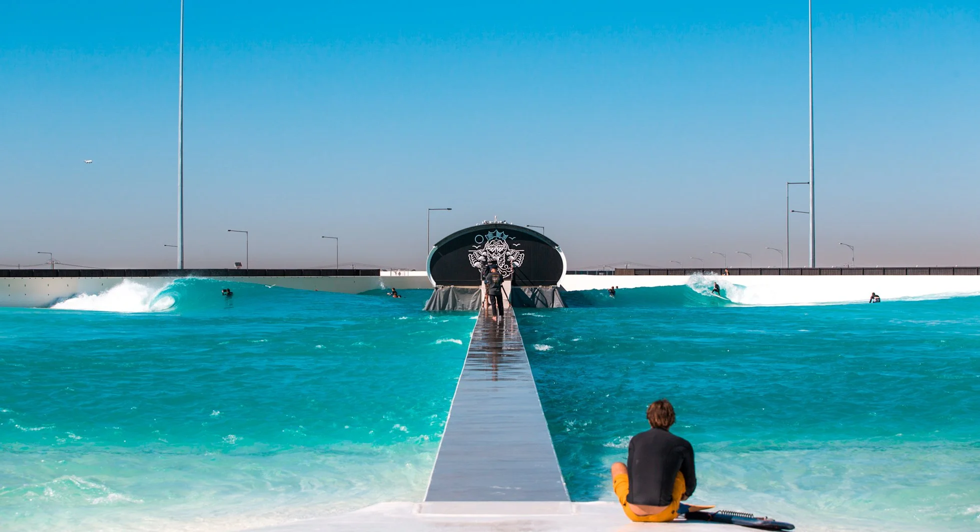 Indoor surf park with artificial wave pool, several surfers, and a walkway leading to a tunnel structure with a lion graphic, under a clear blue sky.