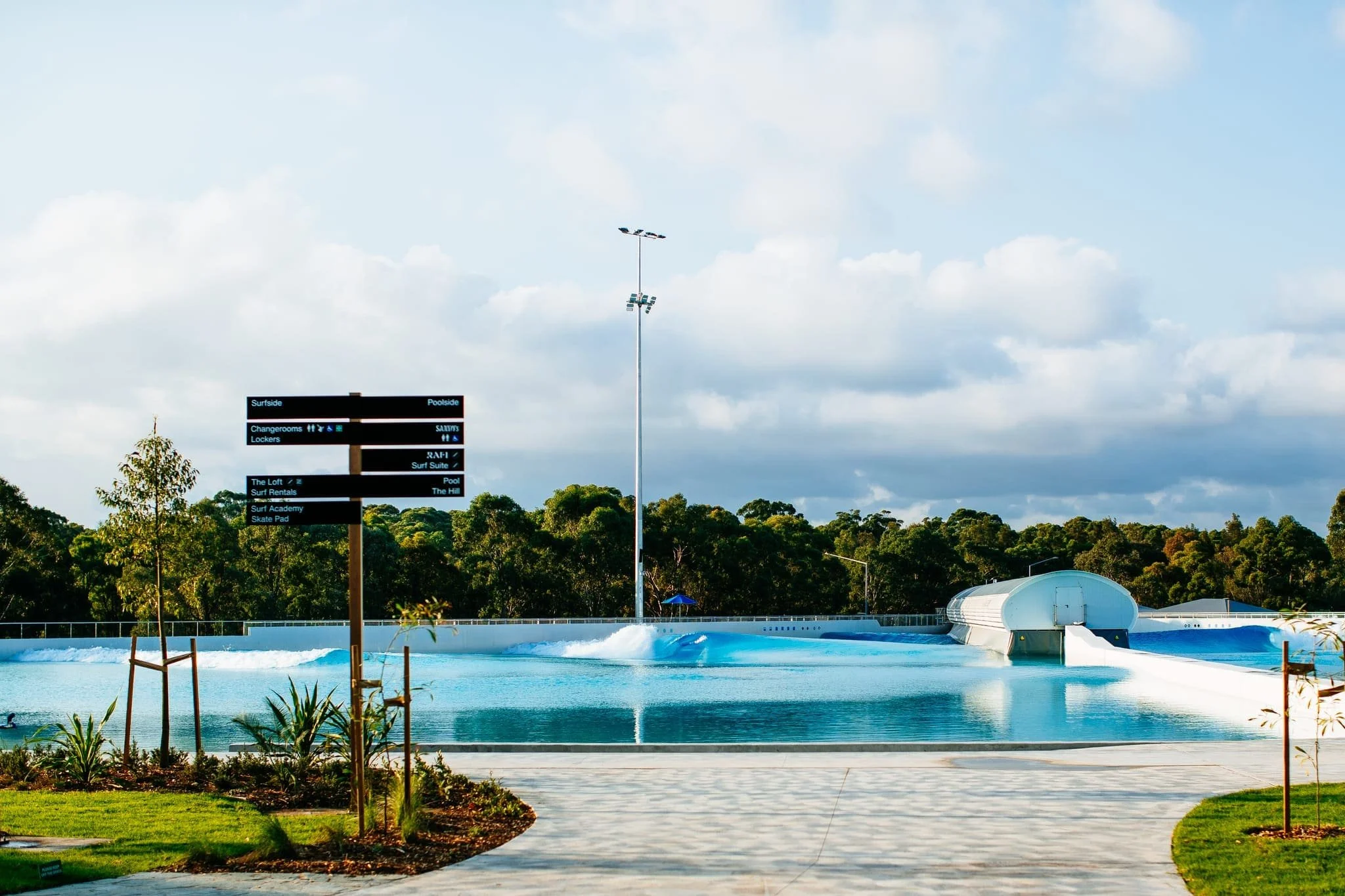 An outdoor water park with a large pool and water slide, surrounded by greenery and trees, with a signpost indicating directions to various park attractions.