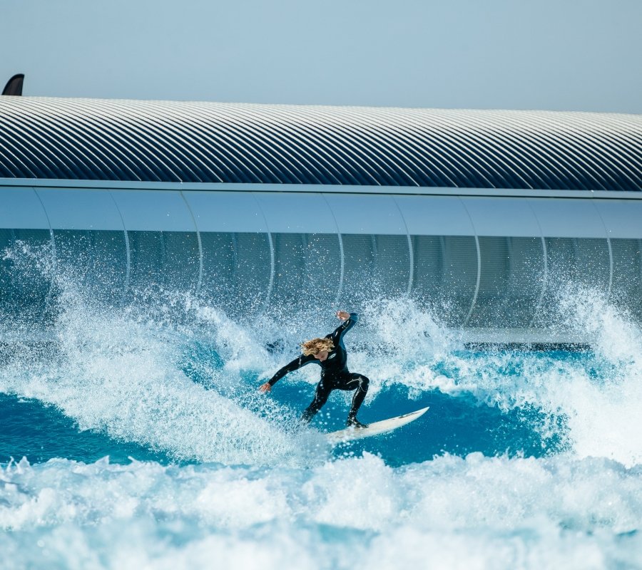 Person in a wetsuit surfing on a wave with a modern building in the background