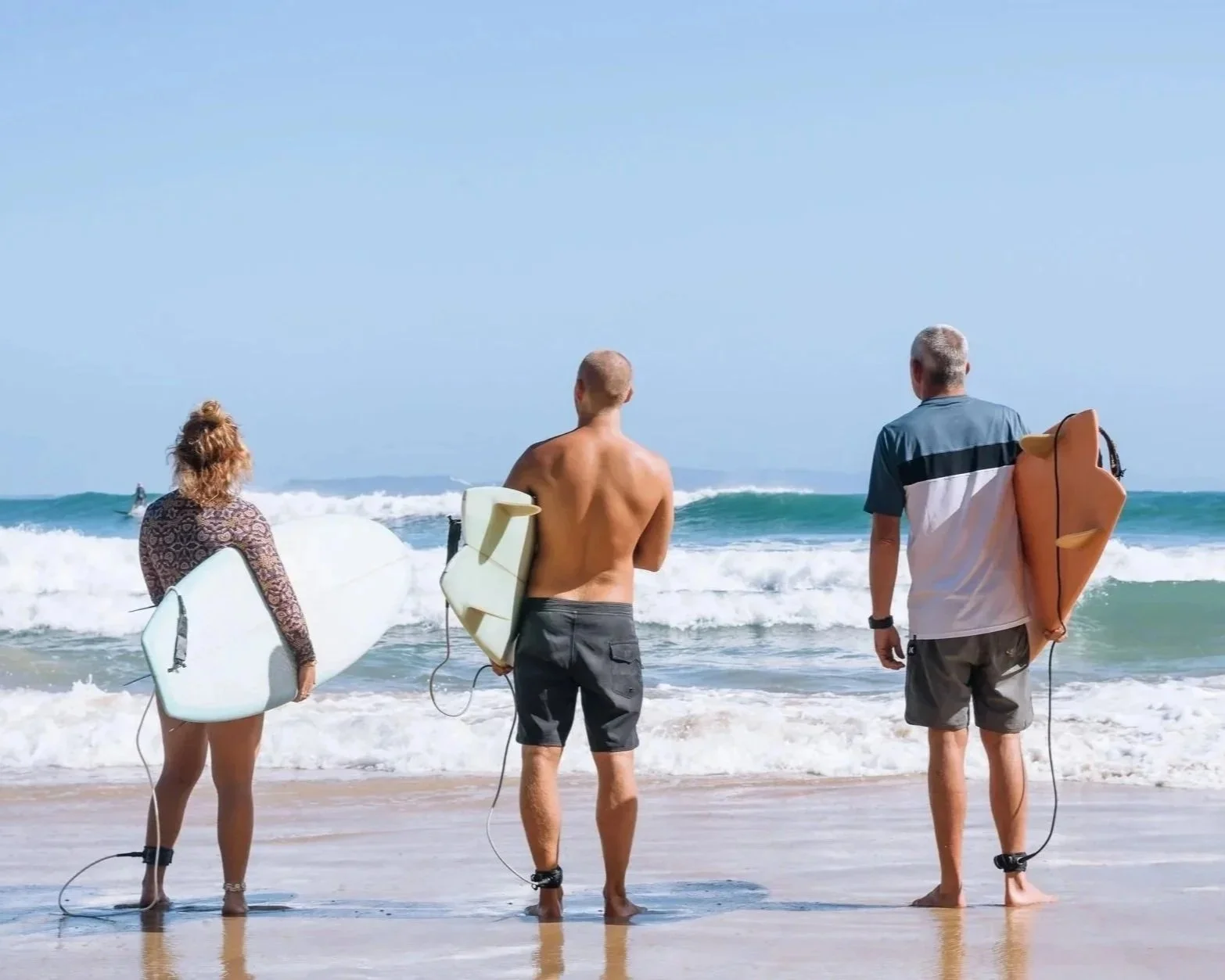 Three surfers with surfboards walking towards the ocean on a sunny beach.