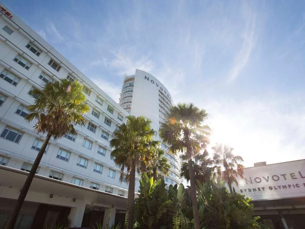 View of the Novotel hotel at Sydney Olympic Park with palm trees in the foreground and sunlight brightening the sky.