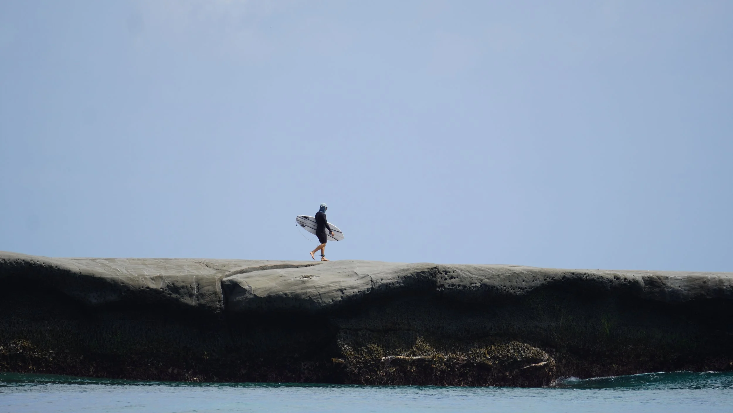 A person holding a surfboard walking along a rocky shoreline under a clear blue sky.