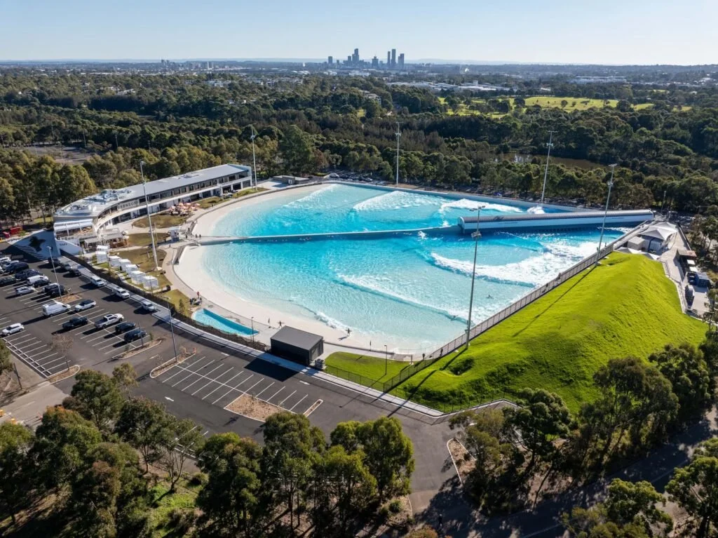 Aerial view of a wave pool with a curved shoreline, surrounded by parking lots and green trees, with a city skyline in the distance.