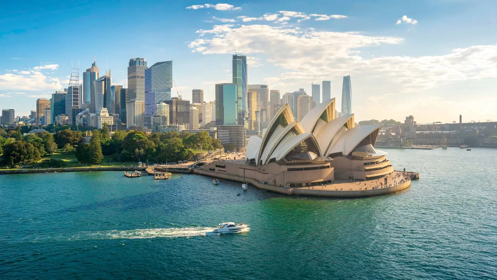 Sydney Opera House with Sydney skyline and harbor, boats on water, sunny day with blue sky and scattered clouds.
