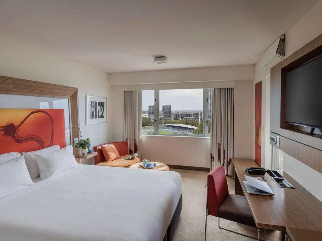 A hotel room with a large bed, a sitting area, a desk with a chair, a wall-mounted TV, and a window showing a cityscape in the background.