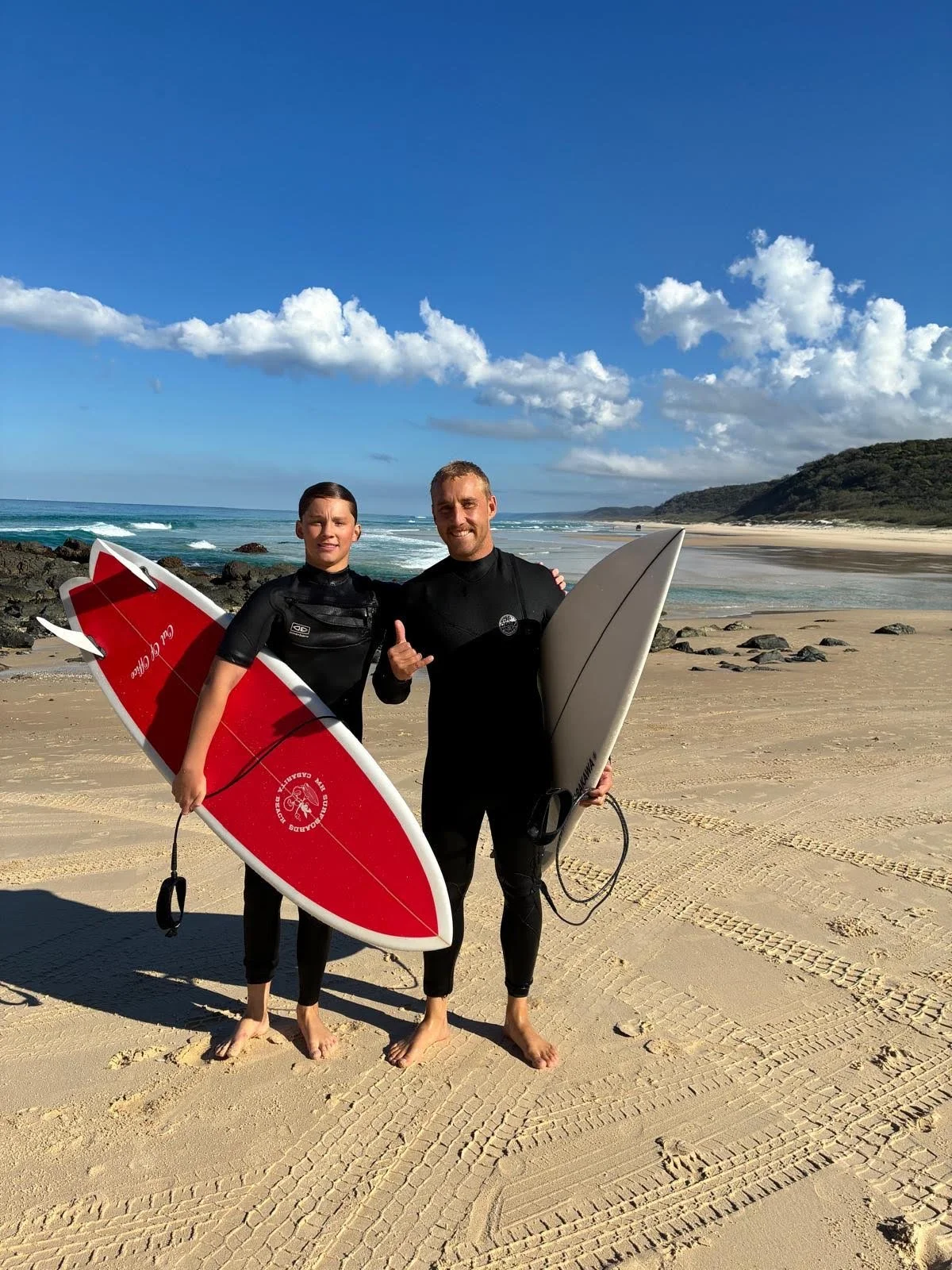 Two surfers in wetsuits standing on a sandy beach, each holding a surfboard, with the ocean, rocks, and hills in the background under a partly cloudy sky.