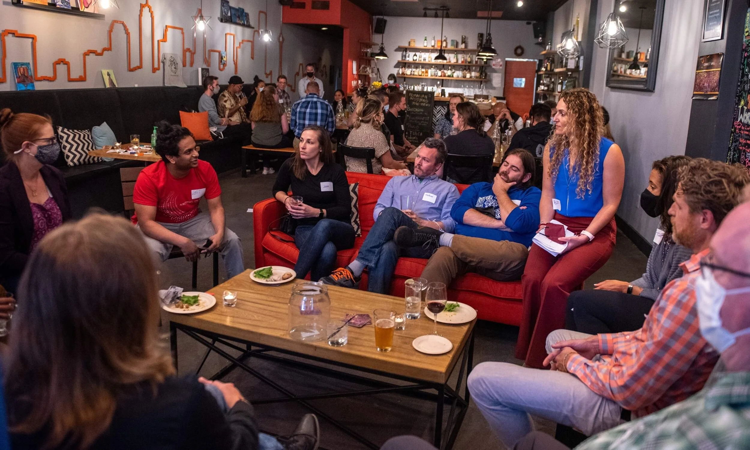 A diverse group of people attending a discussion or social event in a cozy cafe or bar, with some seated on a red sofa and others around a wooden table, engaging in conversation.