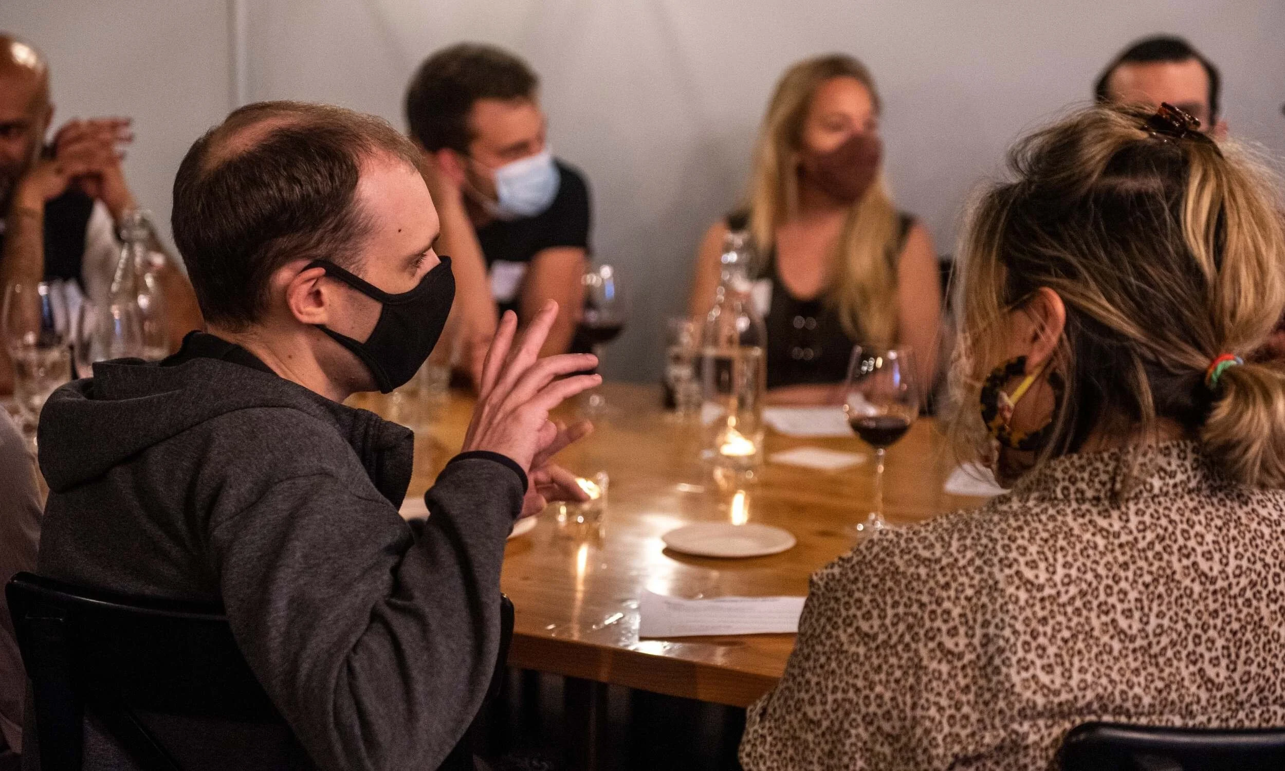 People sitting around a table, wearing masks, engaged in conversation at a social gathering or meeting.